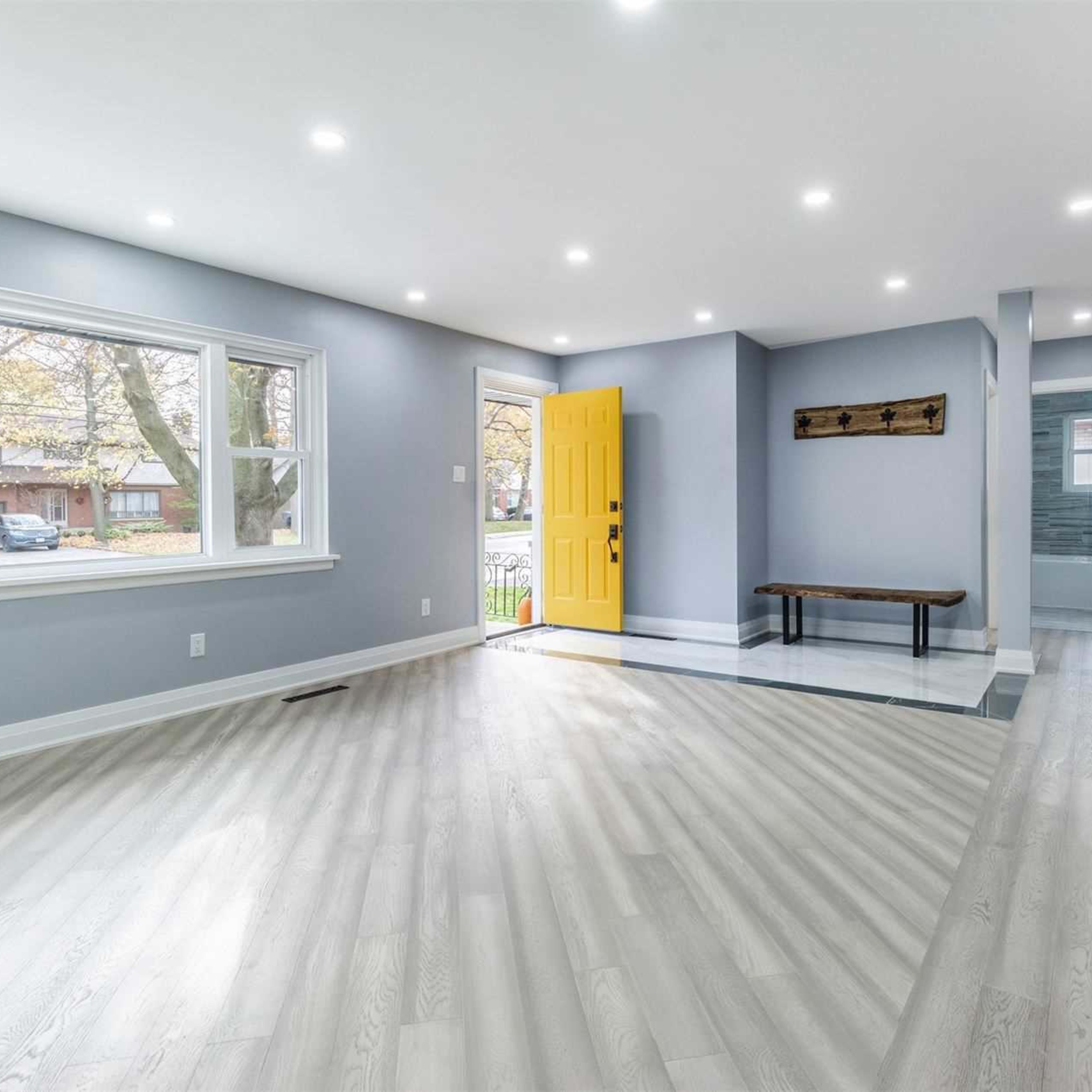 A spacious, well-lit interior room with light gray walls, a yellow front door, and a wooden bench against one wall.