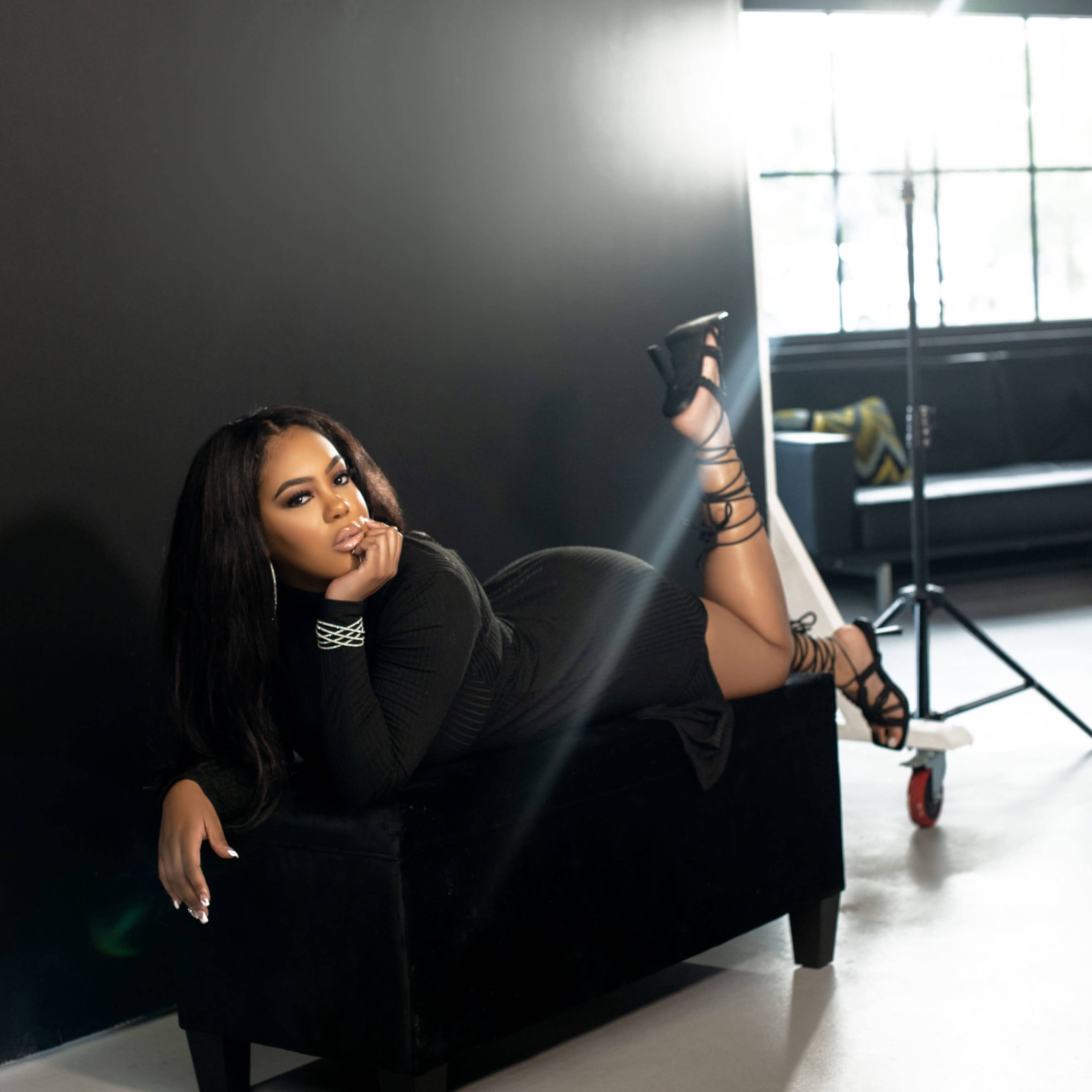 A person in a black dress is lying on a dark bench in a well-lit room with large windows and minimal furnishings.