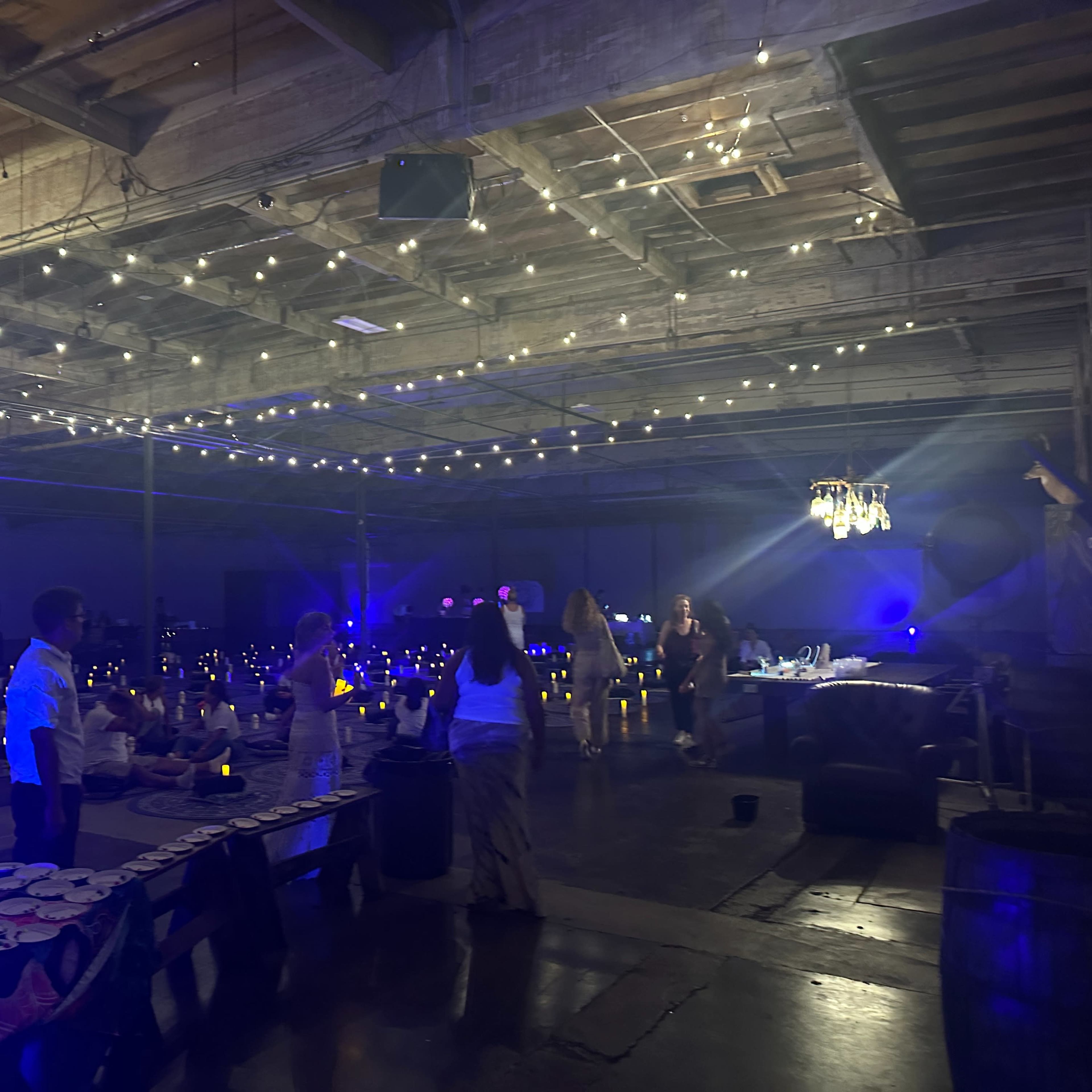A spacious indoor venue with strings of lights overhead, set up for an event with tables arranged and guests mingling under soft blue lighting.
