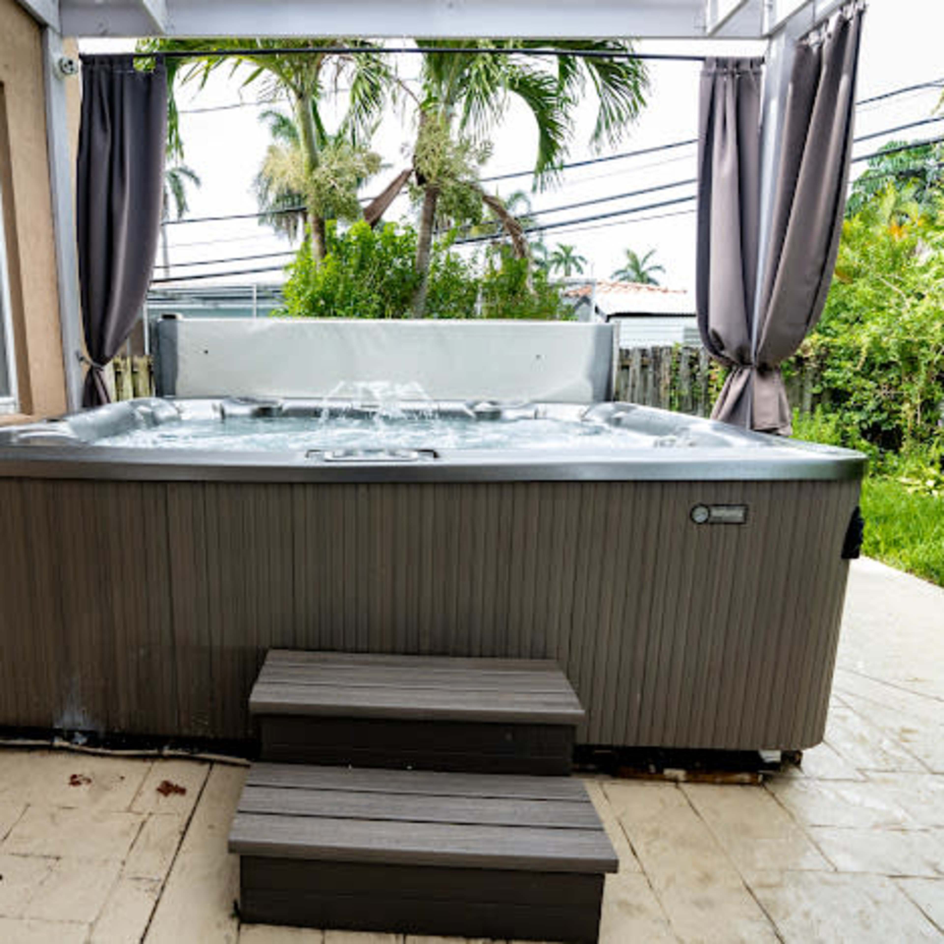 A hot tub sits on a patio next to a house, with steps leading up to it and palm trees visible in the background.