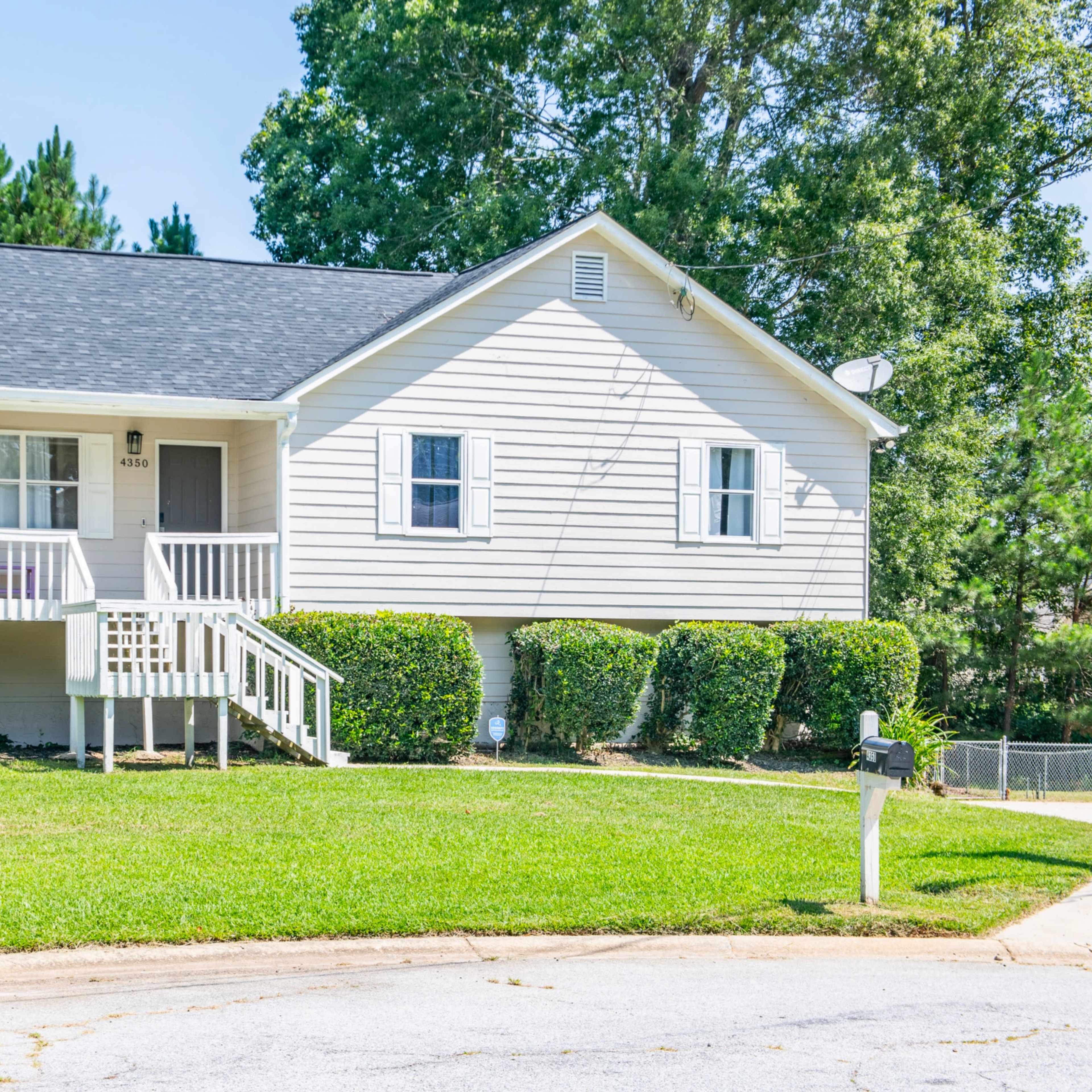 A single-story house with a light gray exterior and a front porch is situated on a green lawn.