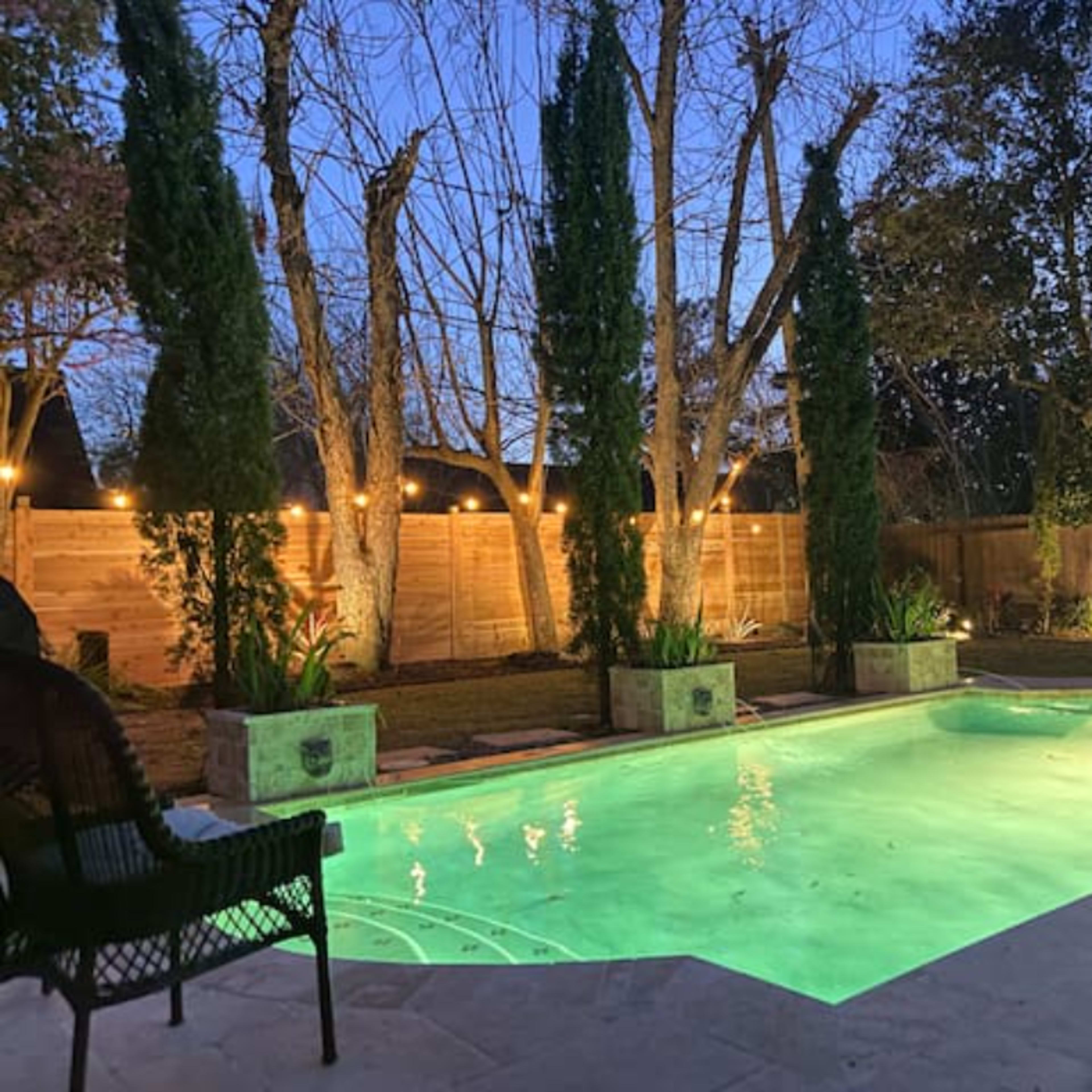 A well-lit backyard features a swimming pool surrounded by greenery and trees at dusk.