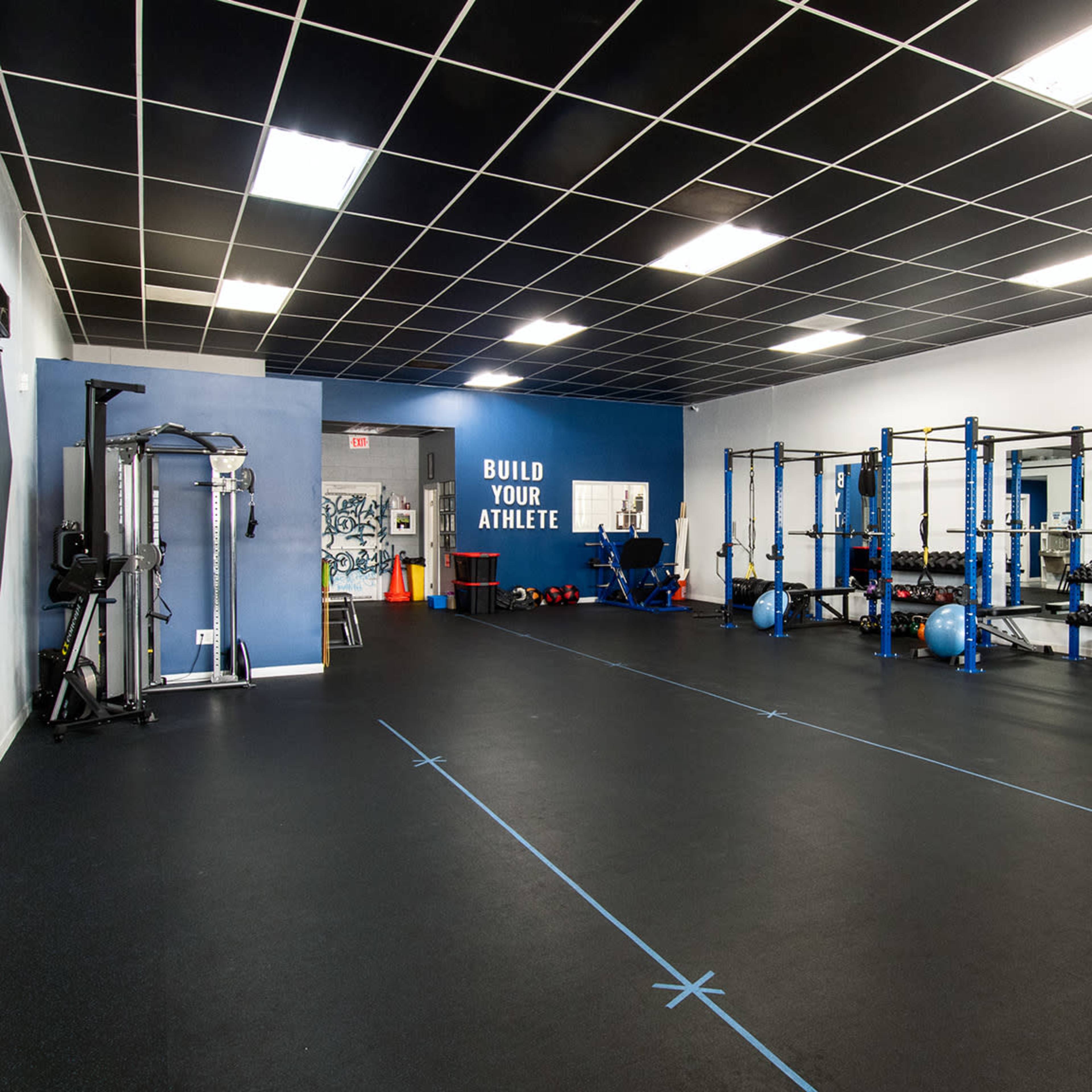A spacious gym interior featuring weightlifting equipment, blue walls, and a large sign that reads "BUILD YOUR ATHLETE."