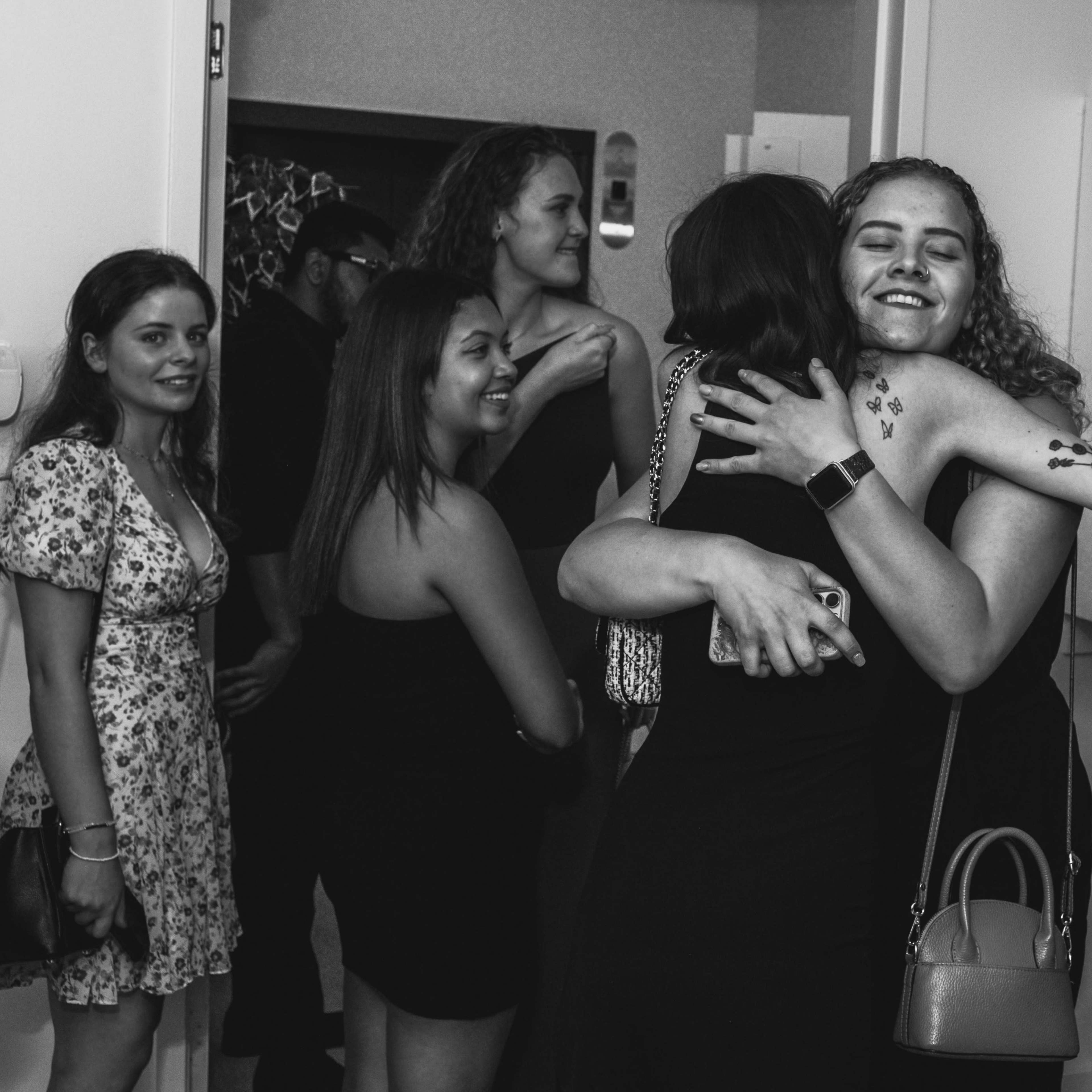 A group of young adults is gathered in a doorway, with two women embracing while others look on.