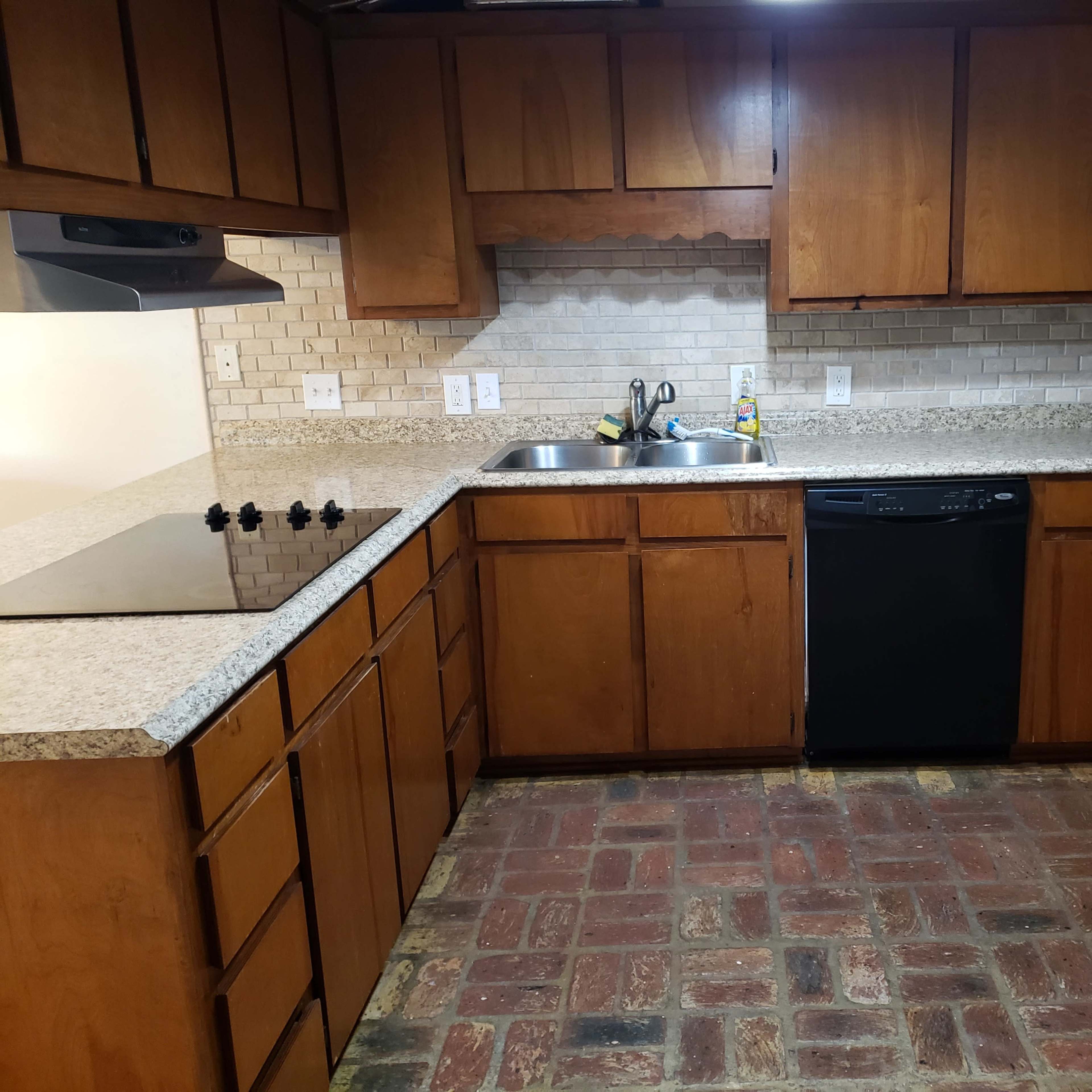 A kitchen with wooden cabinets, a granite countertop, a sink, a dishwasher, and a brick-patterned floor.