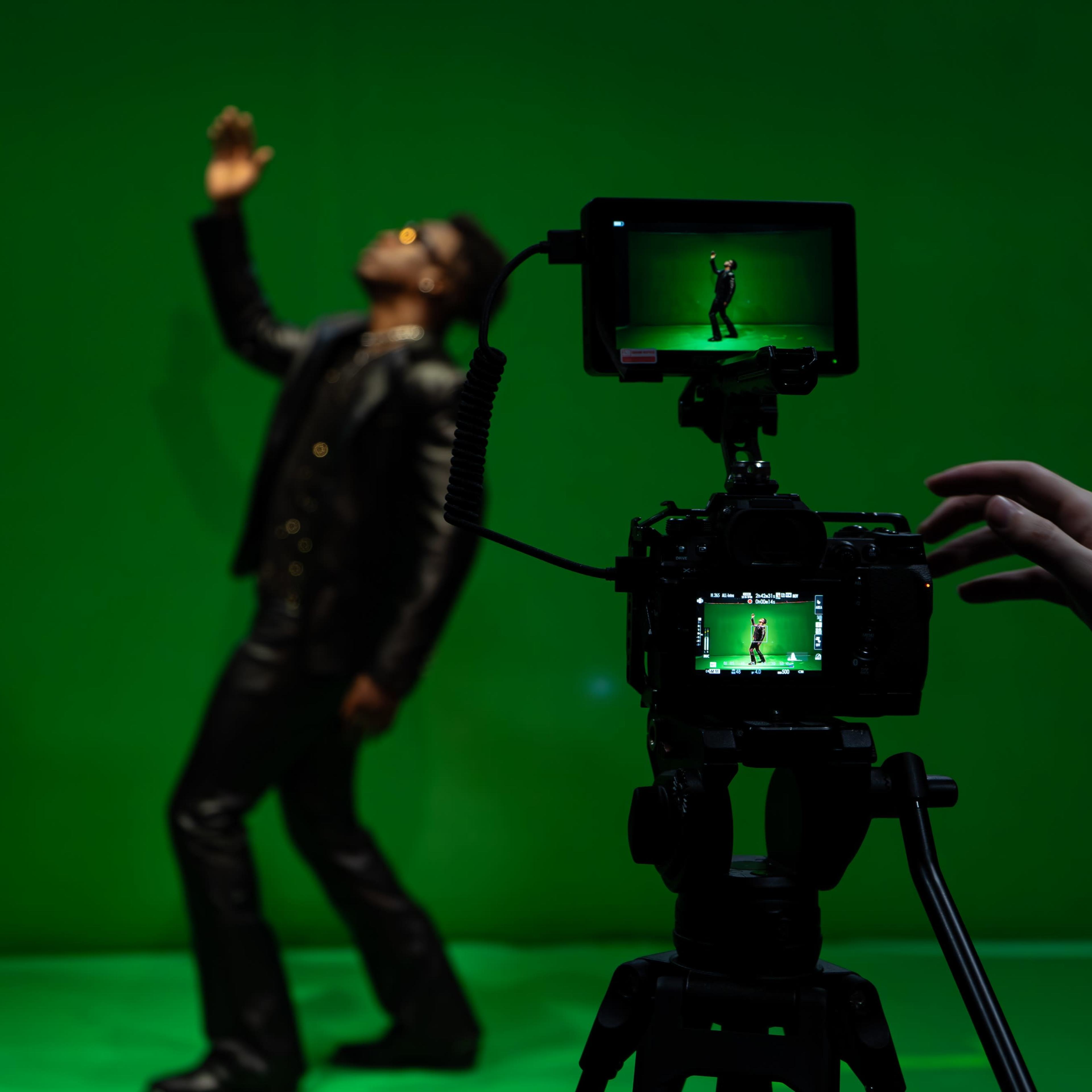 A person in a black outfit gestures dramatically in front of a green screen while being filmed by a camera on a tripod.