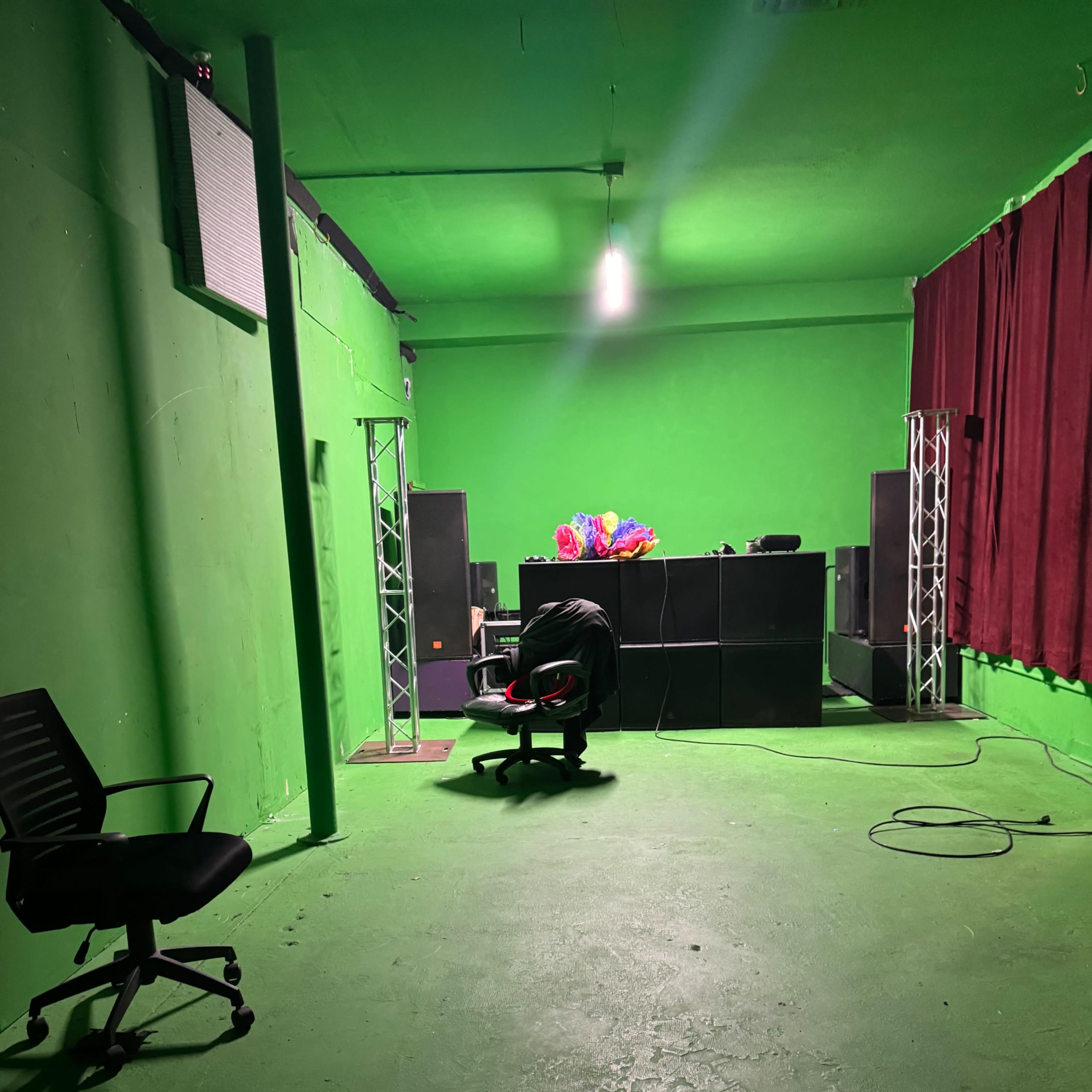 An empty room with green walls and floor, featuring a black office chair, a second black chair, and a sound system against the wall.