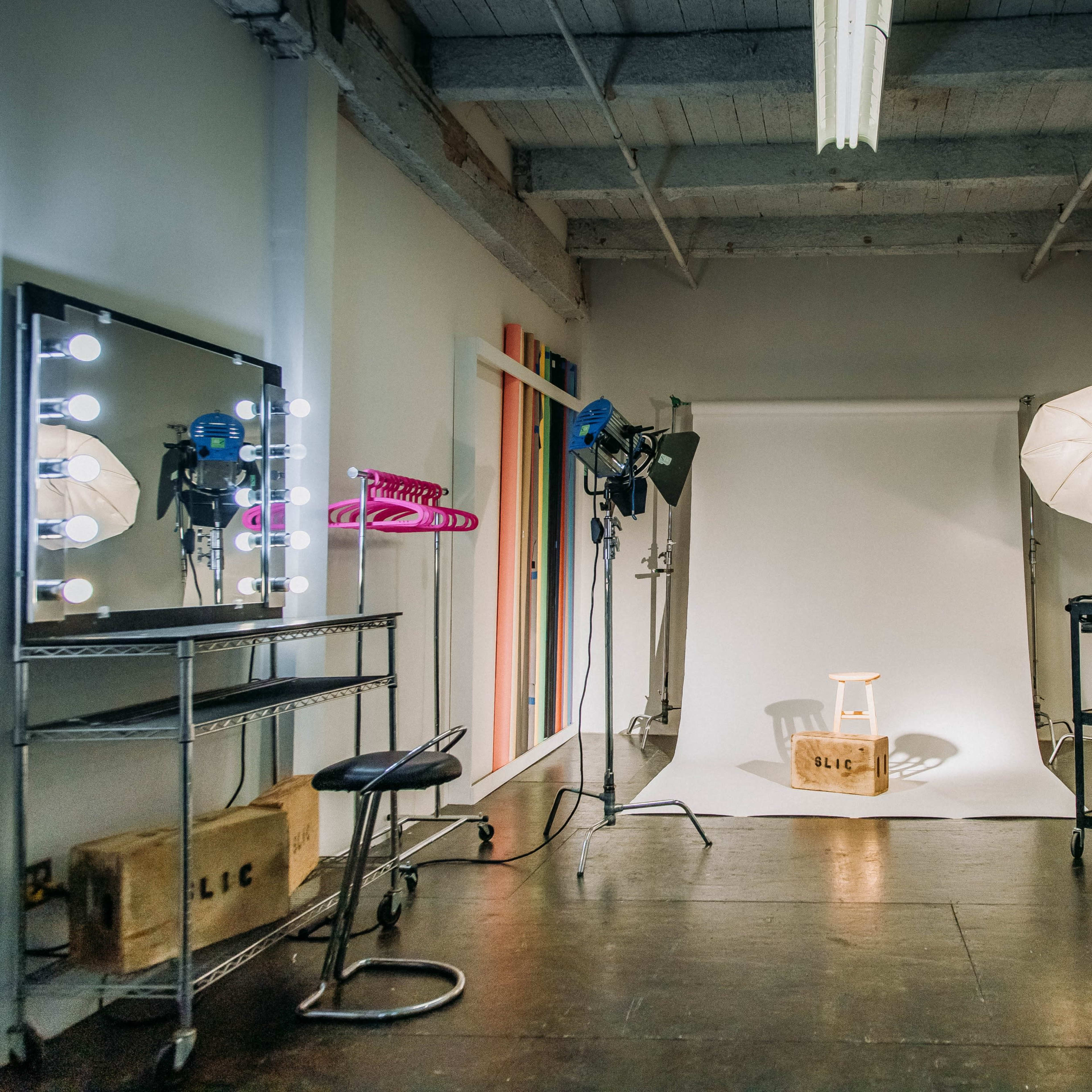 A photography studio features a light backdrop, a chair on a stand, and various lighting equipment arranged around the space.