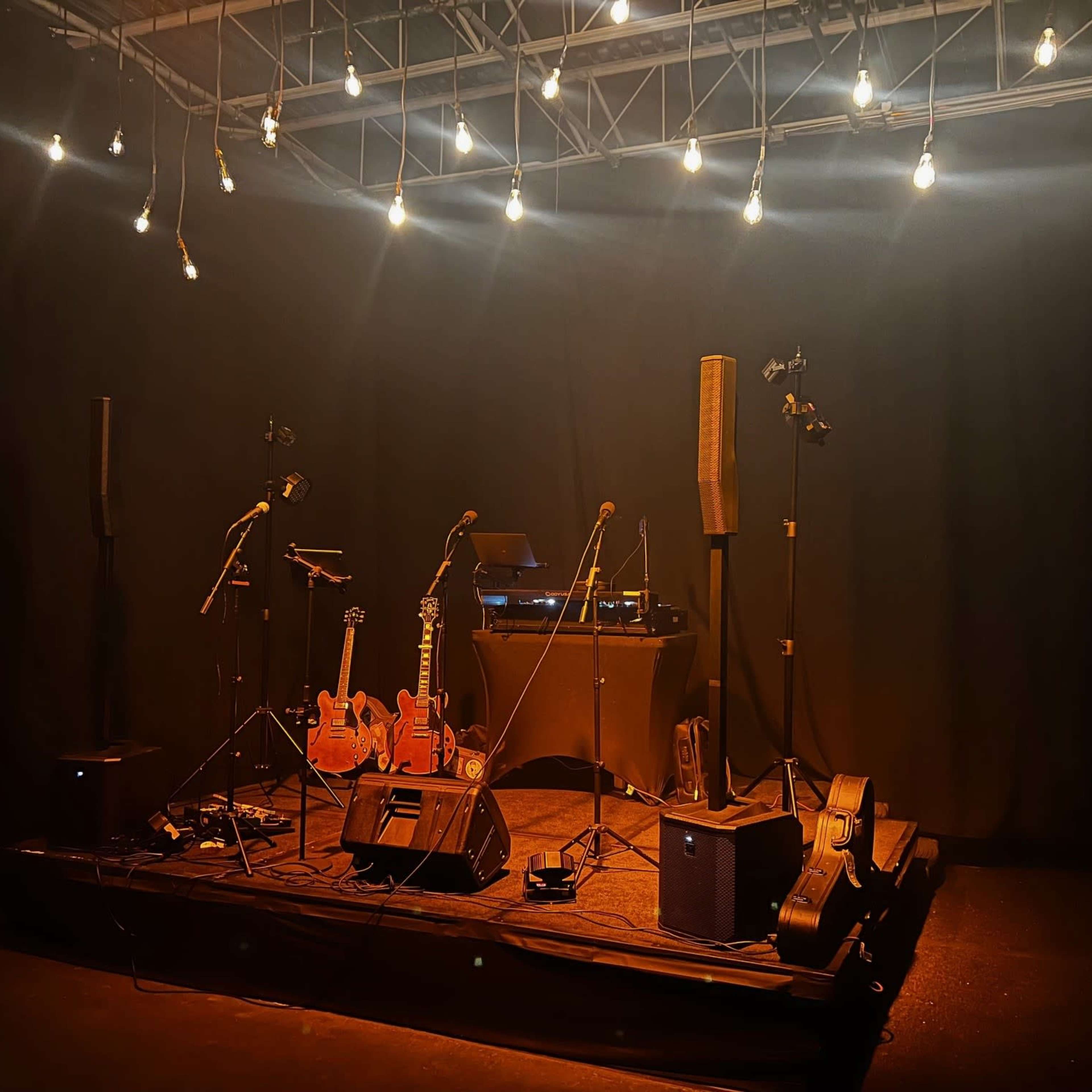 A dimly lit stage set up with guitars, microphones, and sound equipment surrounded by hanging lights.