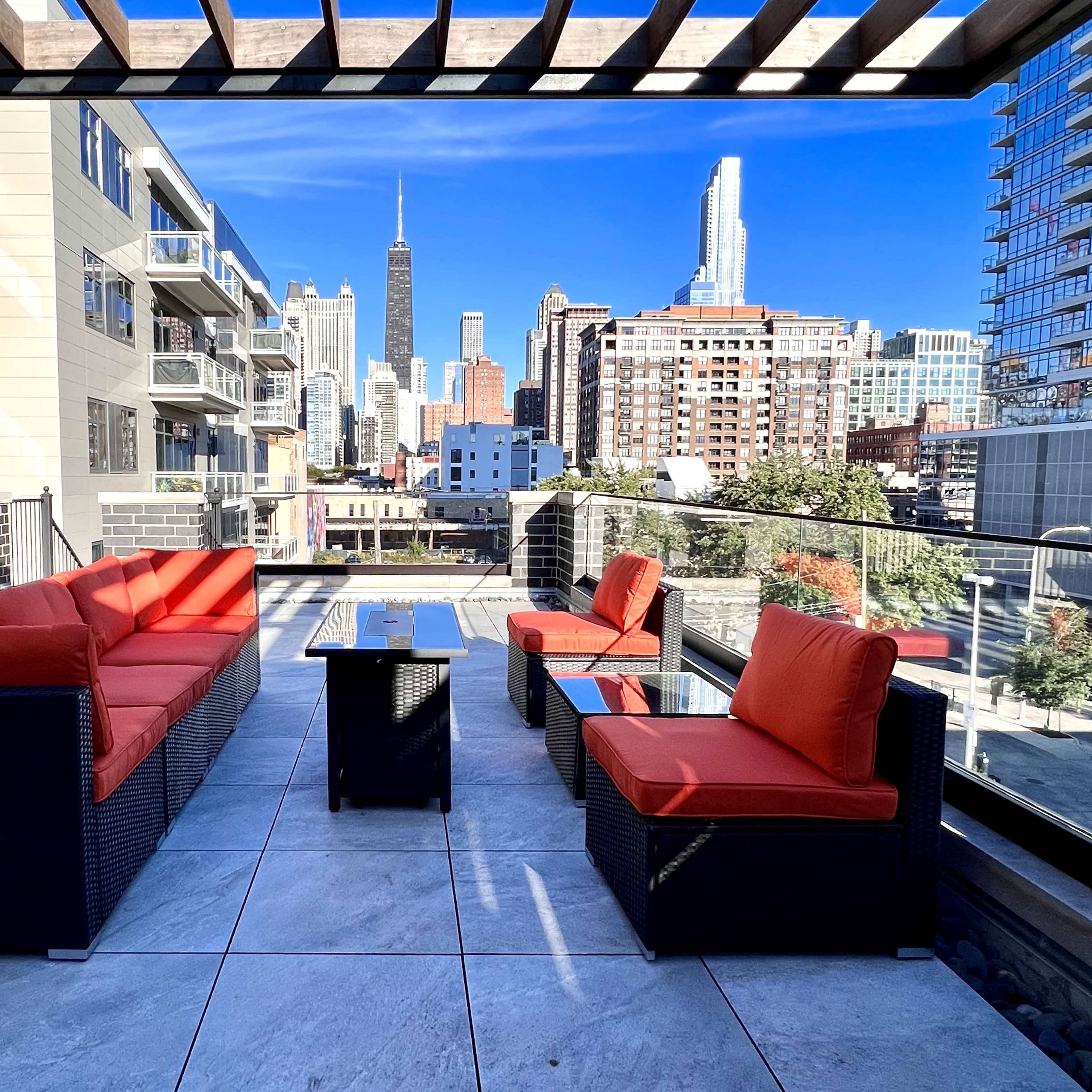 A rooftop terrace features orange cushion seating and a table, overlooking a city skyline with skyscrapers, including the Empire State Building.