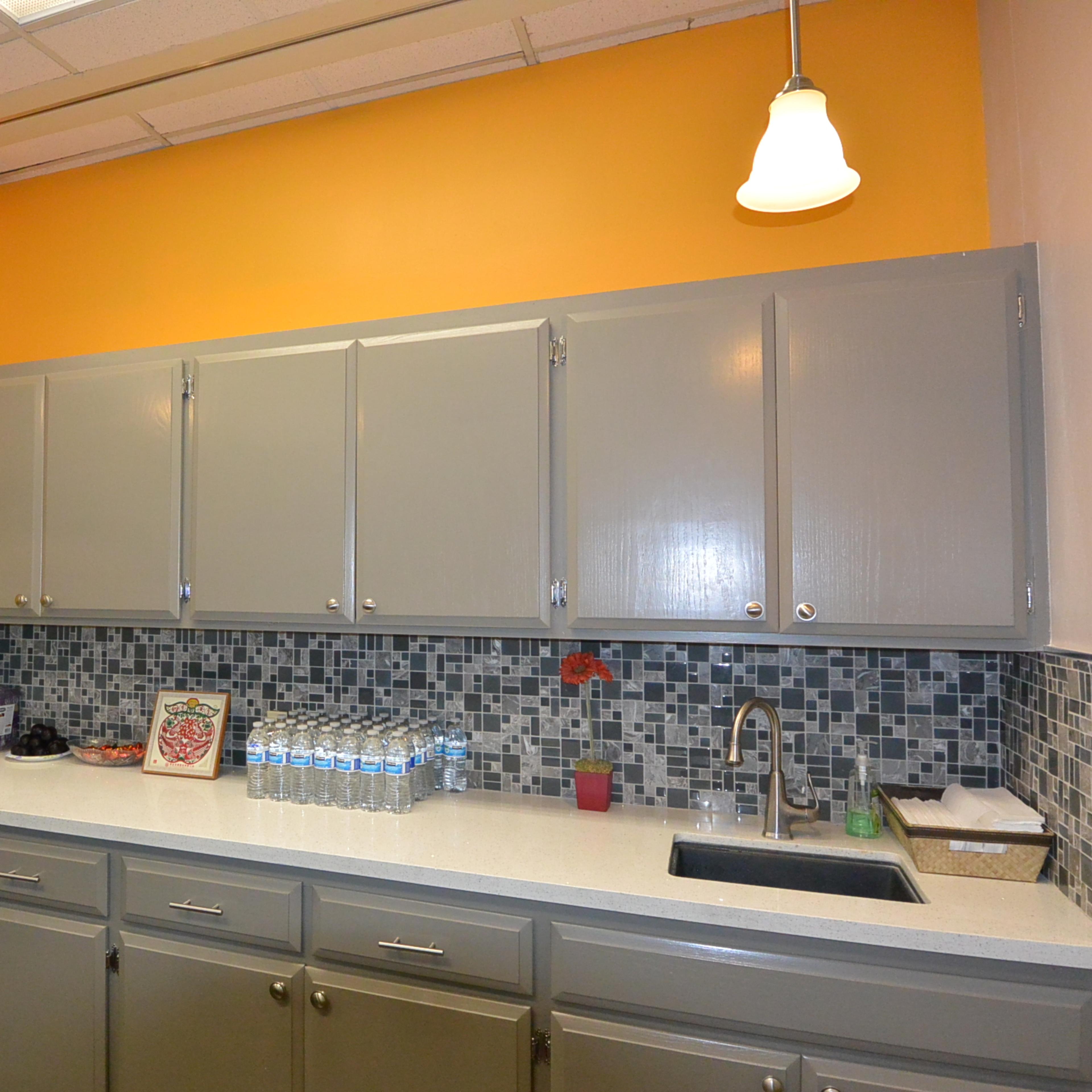 A kitchen with gray cabinets, a mosaic tile backsplash, a white countertop, and orange walls, illuminated by pendant lights.