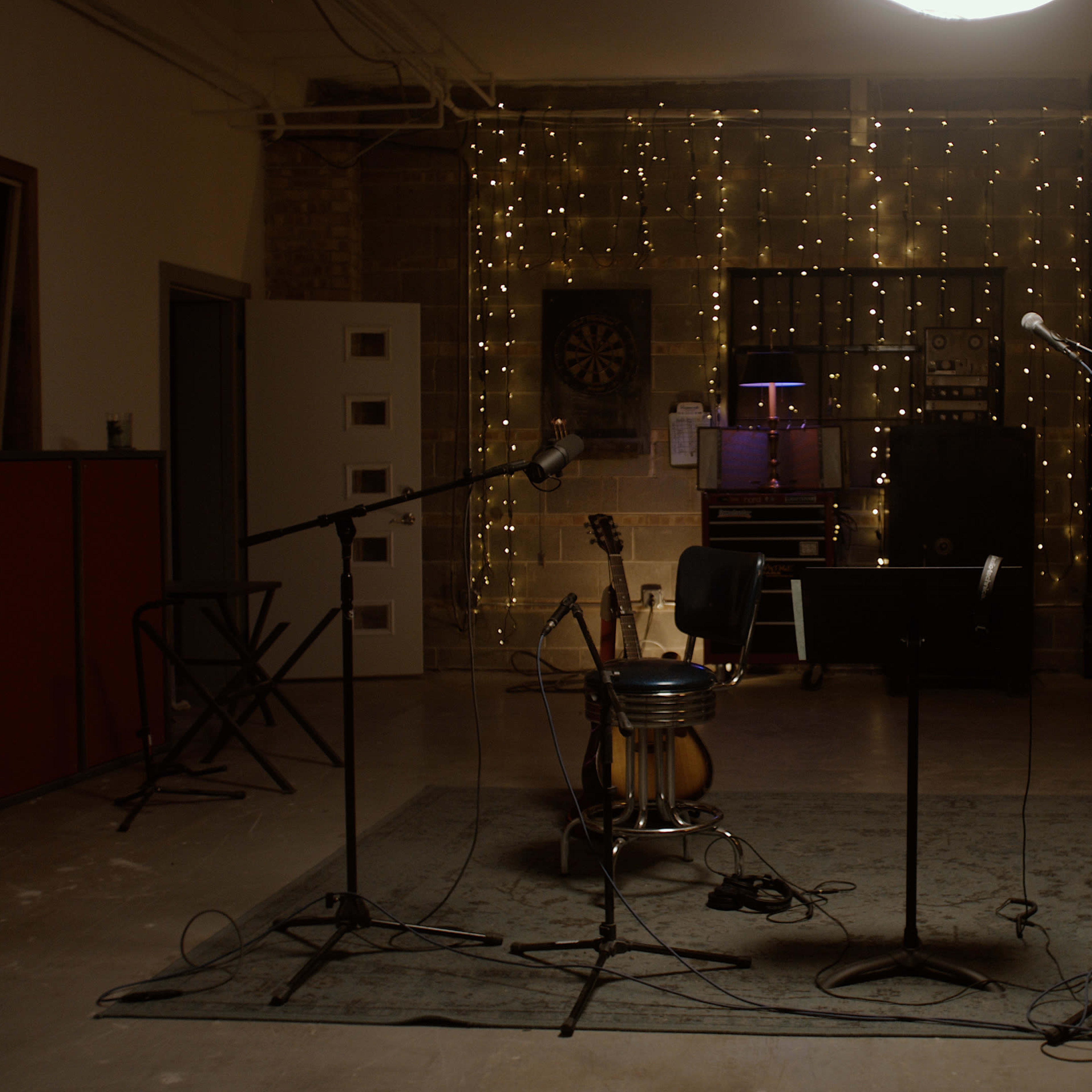 A recording studio with microphones set up on a rug, an acoustic guitar leaning against a chair, and string lights illuminating the background.