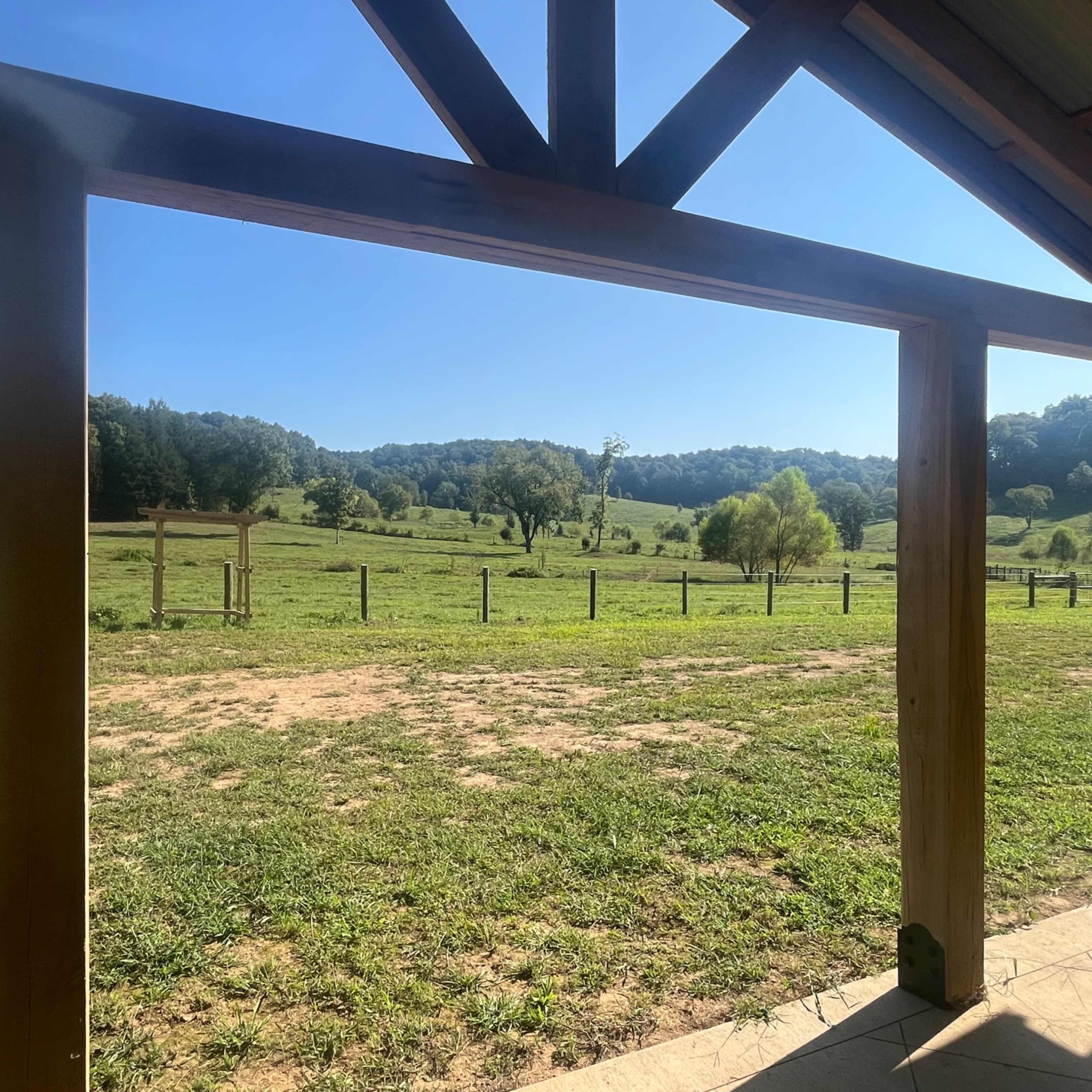 A wide open field framed by wooden beams, with rolling hills and trees in the background under a clear sky.