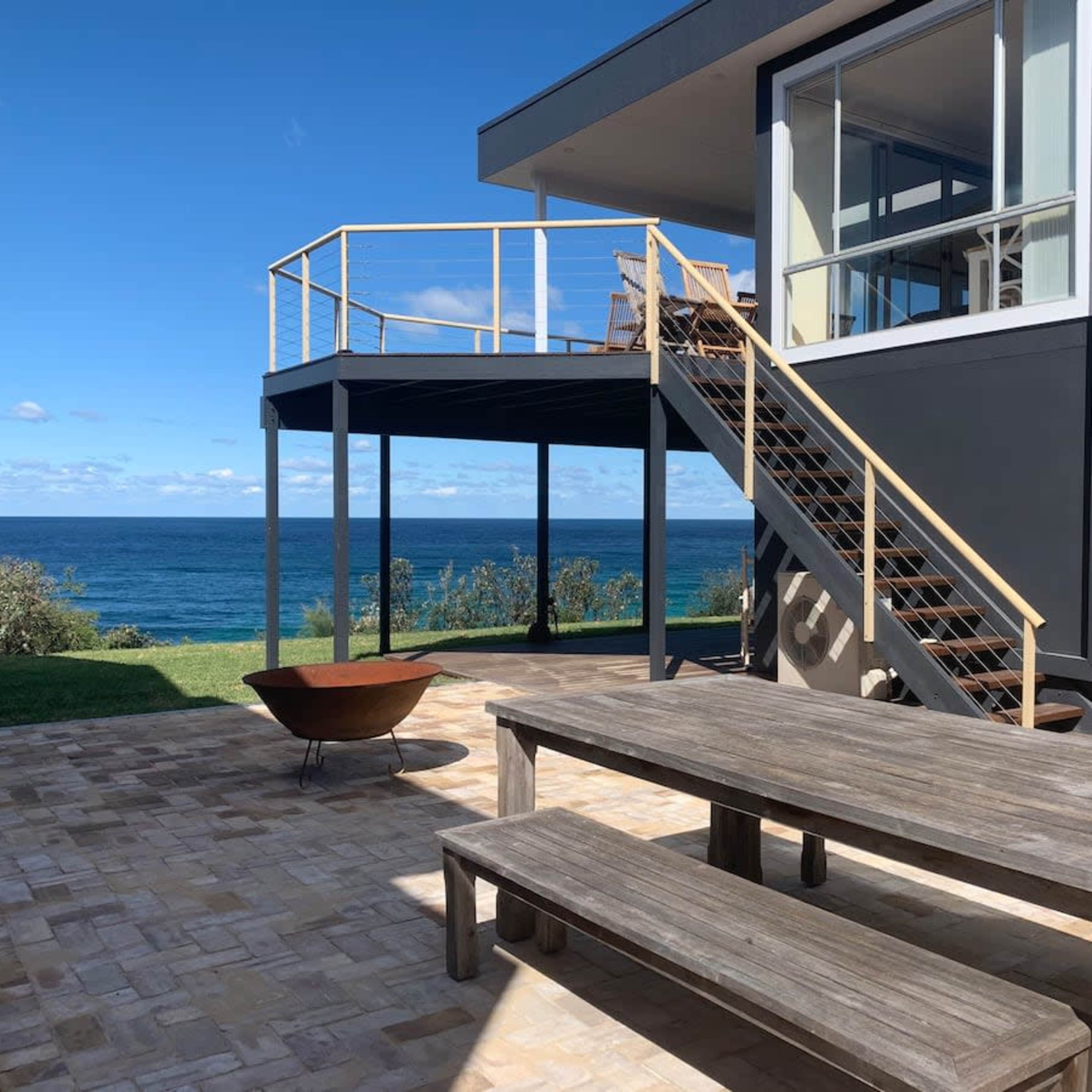 A wooden table and benches are set on a stone patio next to a modern house with a staircase leading to an upper deck and views of the ocean.