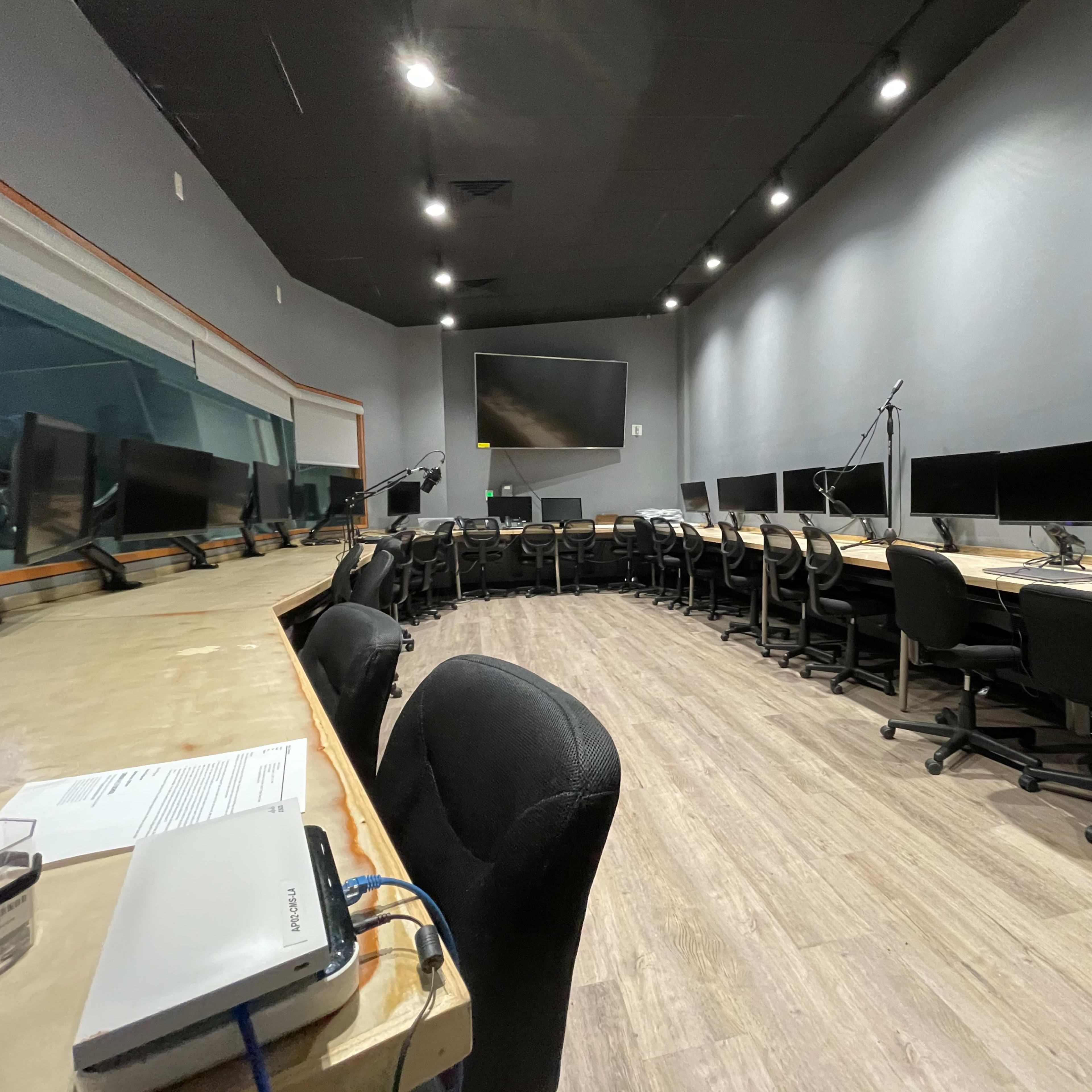 A modern recording studio with a circular arrangement of desks and monitors, equipped with chairs and a large screen at the front.