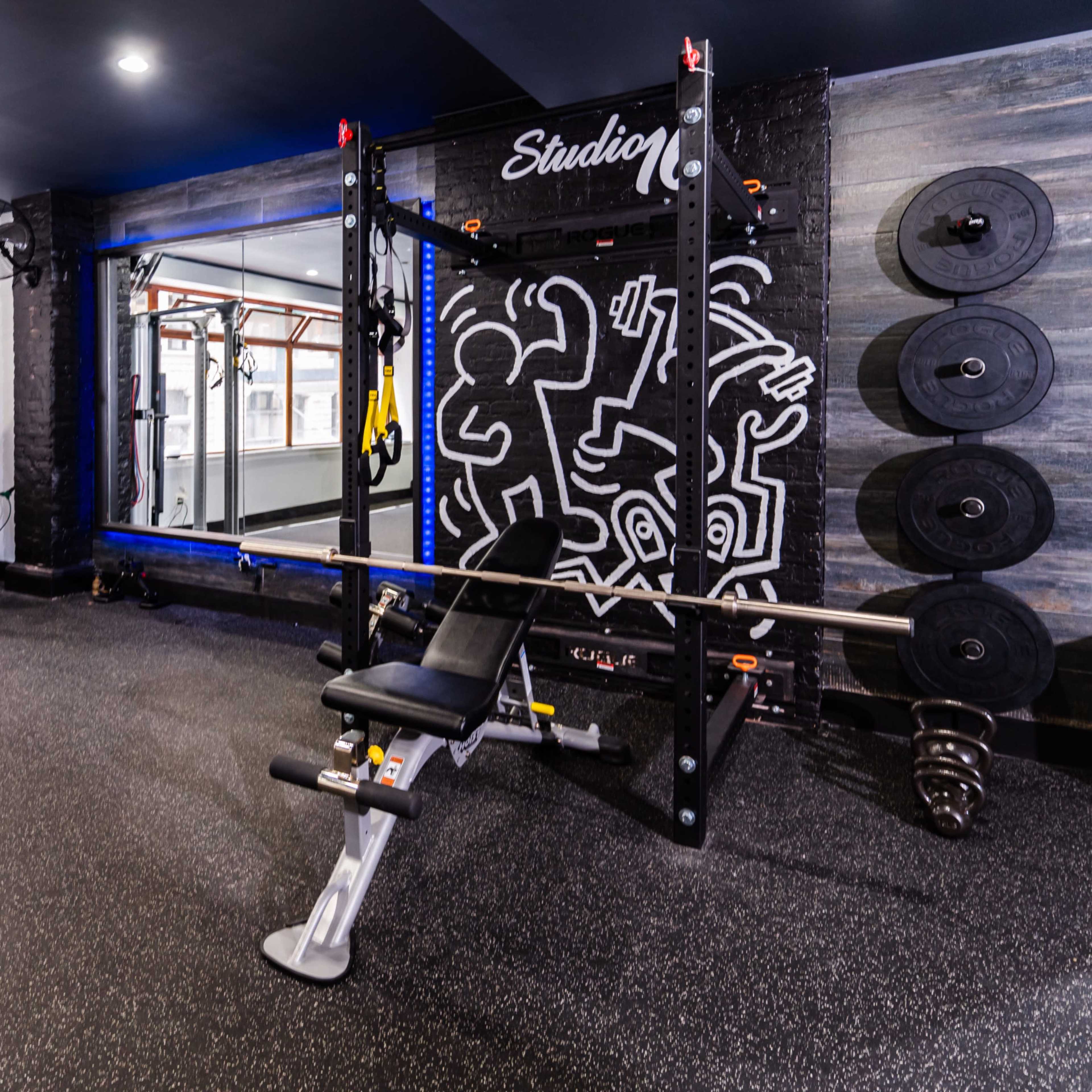 A well-equipped home gym featuring a weight bench, exercise machine, and free weights against a colorful wall with artwork.