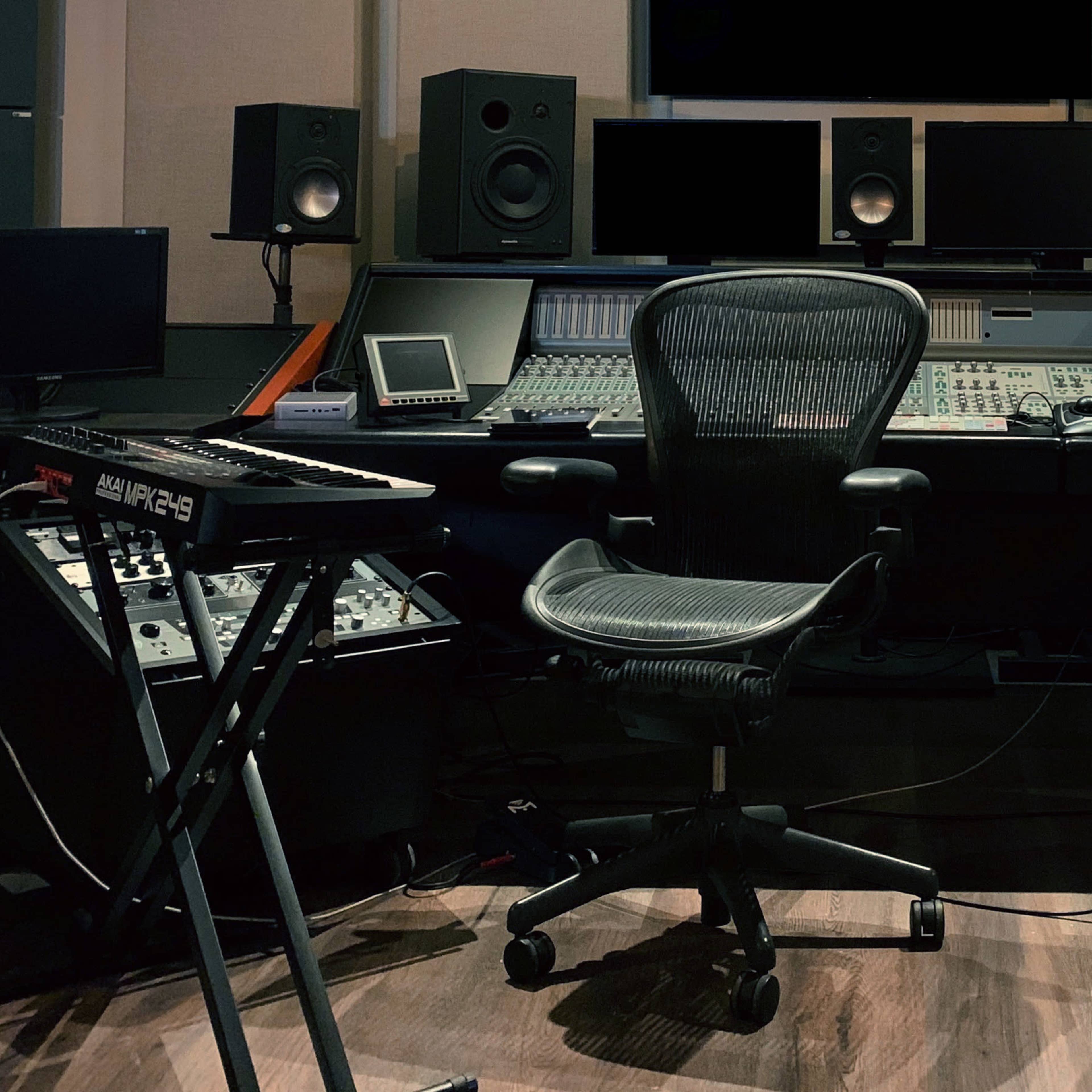 A music production studio with a central mixing console, multiple monitors, speakers, and various music equipment arranged around an ergonomic office chair.