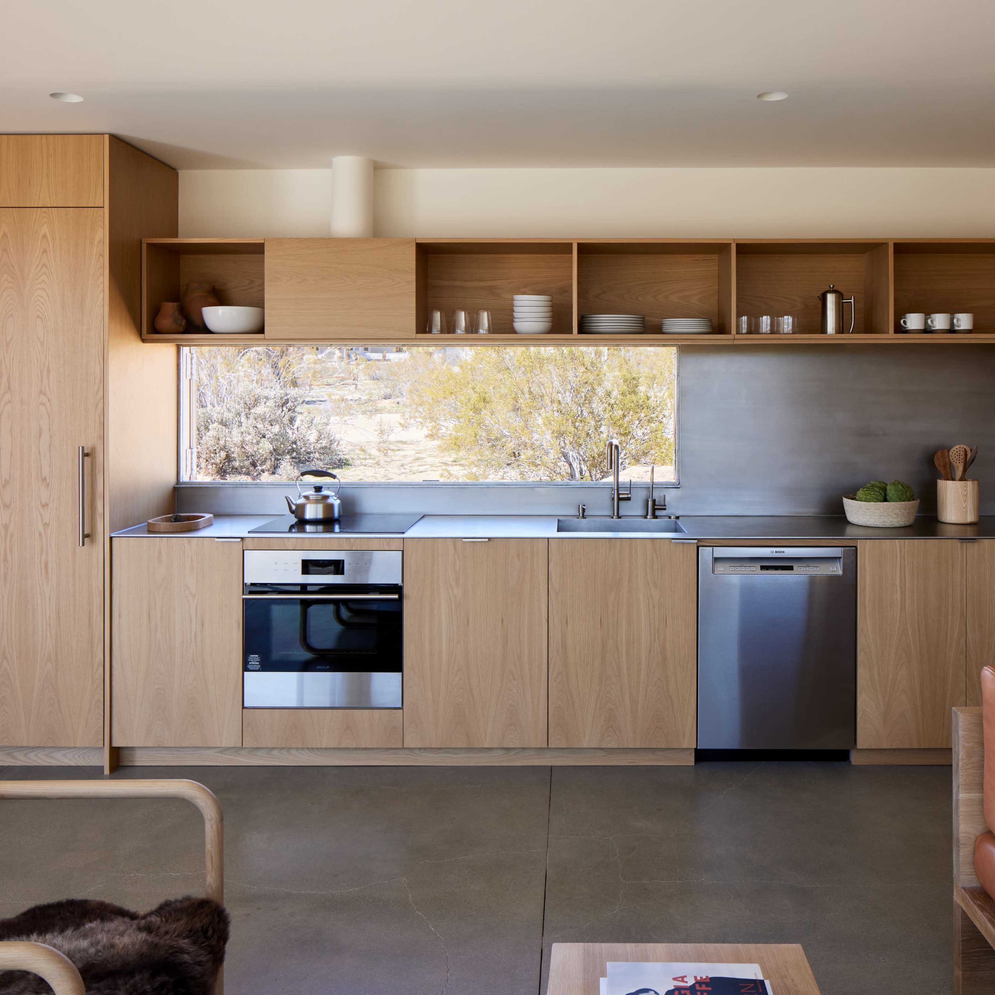 A modern kitchen with wooden cabinetry, stainless steel appliances, and a large window overlooking a natural landscape.
