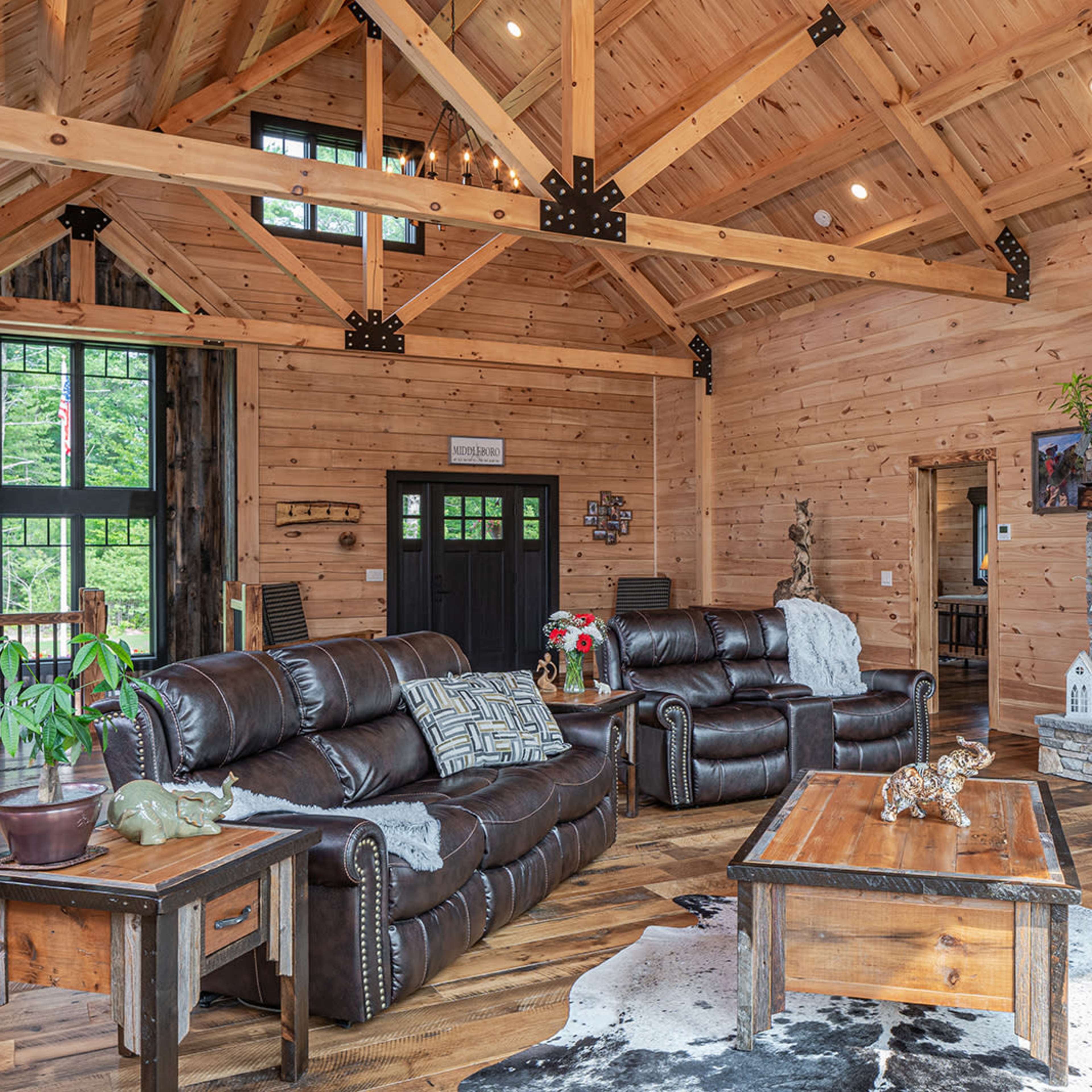 A spacious living room featuring wooden beams, a stone fireplace, and leather seating arranged around a central coffee table on a wooden floor.