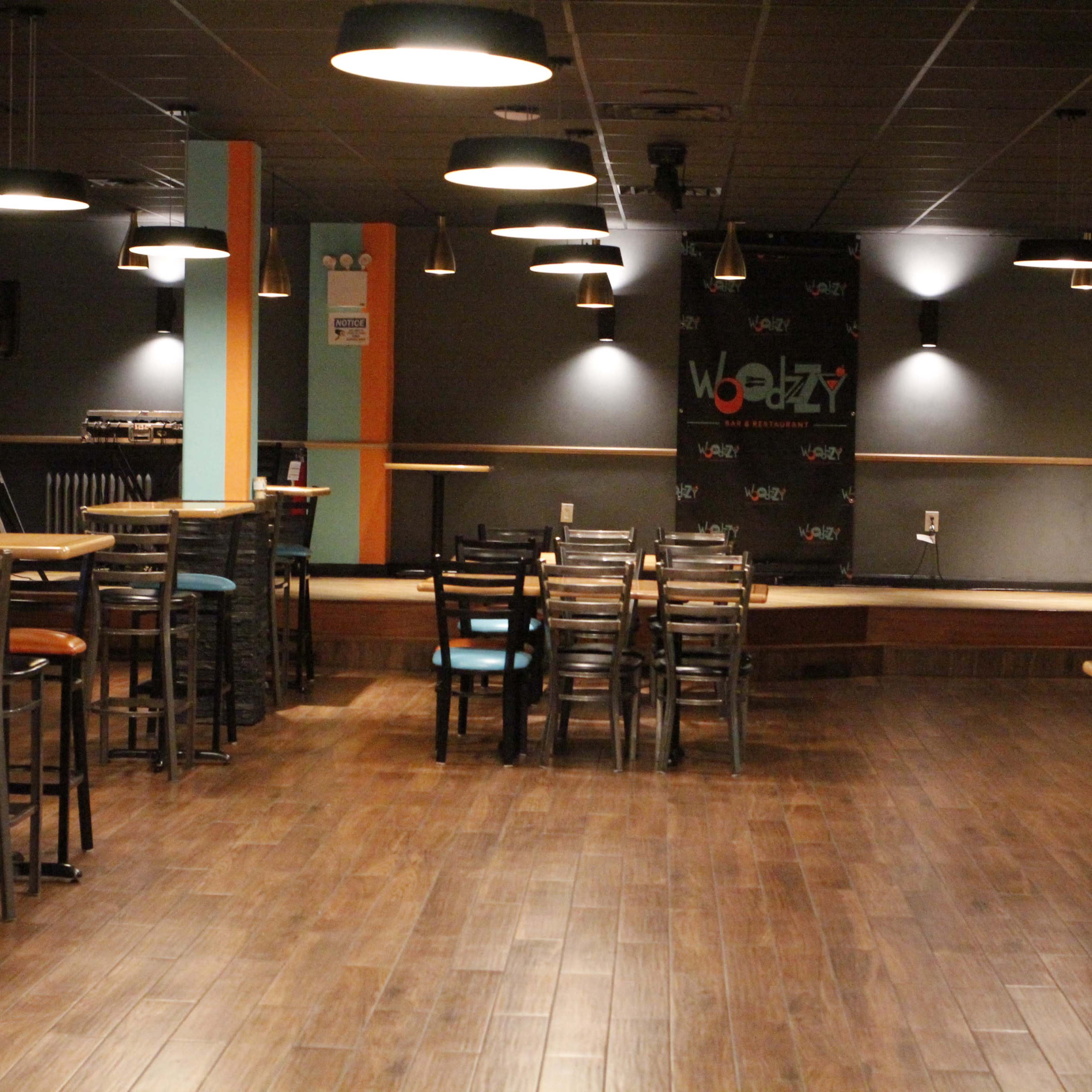 A dimly lit café or bar with wooden flooring, empty tables and chairs, and a small stage at the back with a microphone setup.
