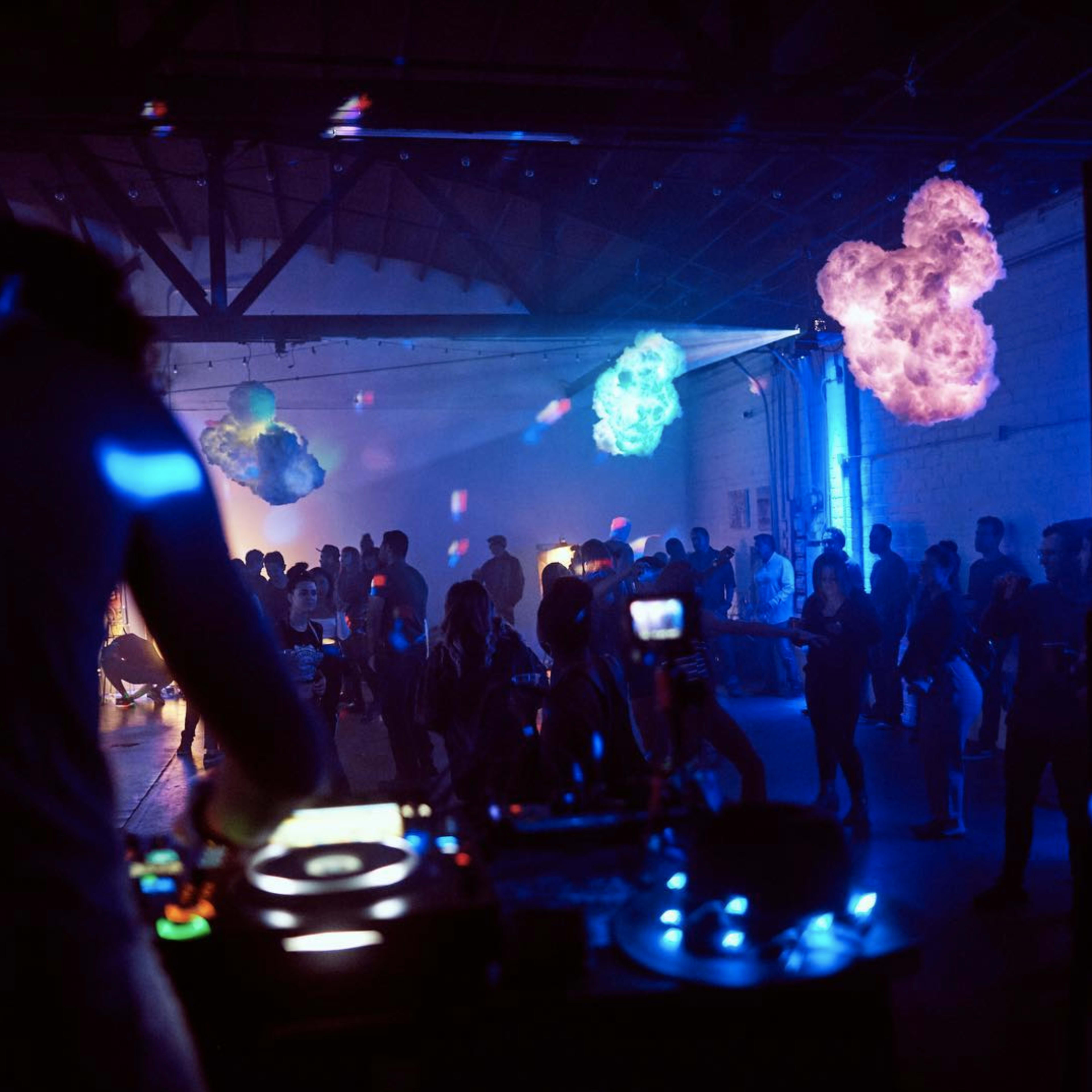 A DJ performs at a dimly lit party featuring colorful lighting effects and a crowd mingling in a spacious venue.