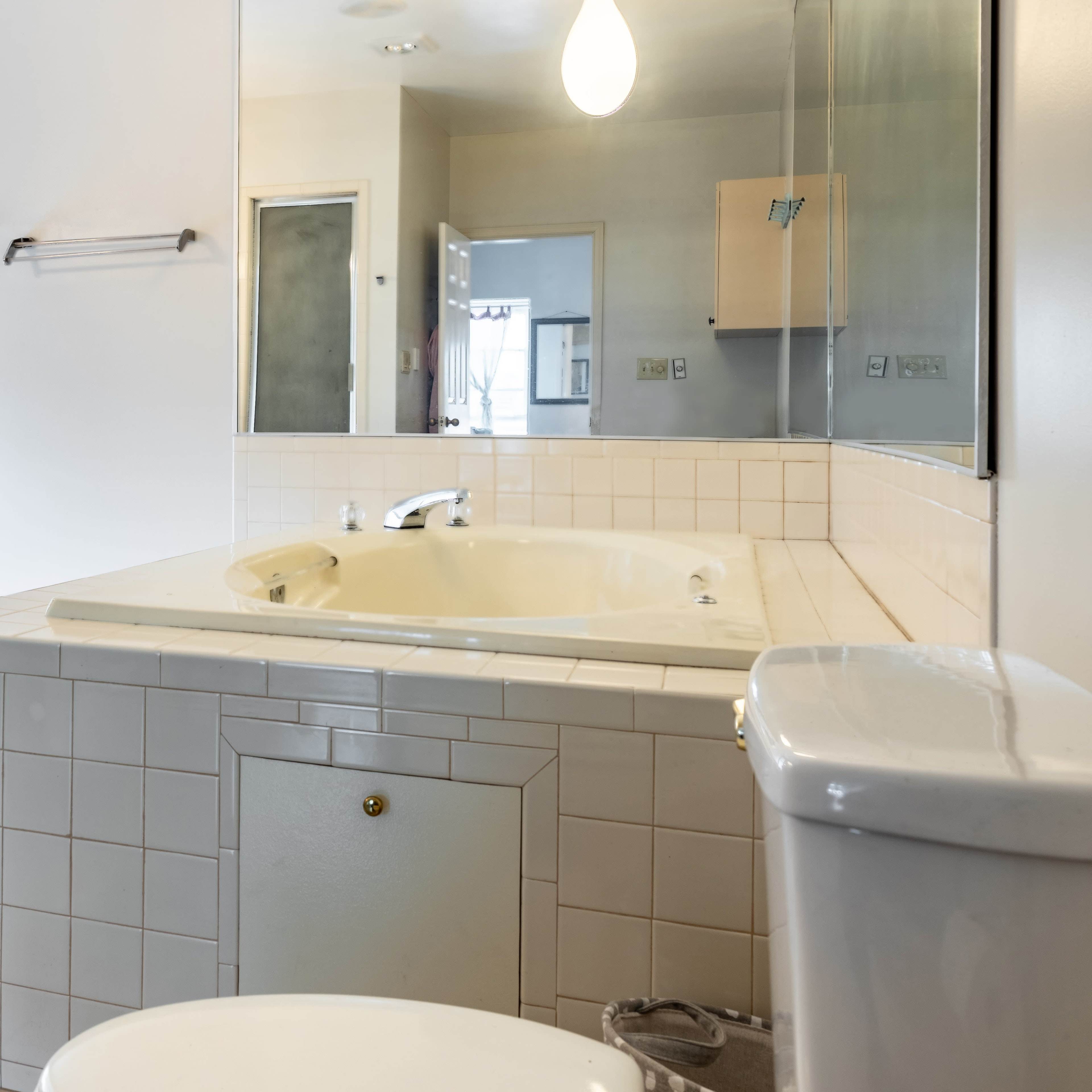 A bathroom features a large tub, a toilet, a sink with cabinets, and a mirror, with natural light coming through a window.