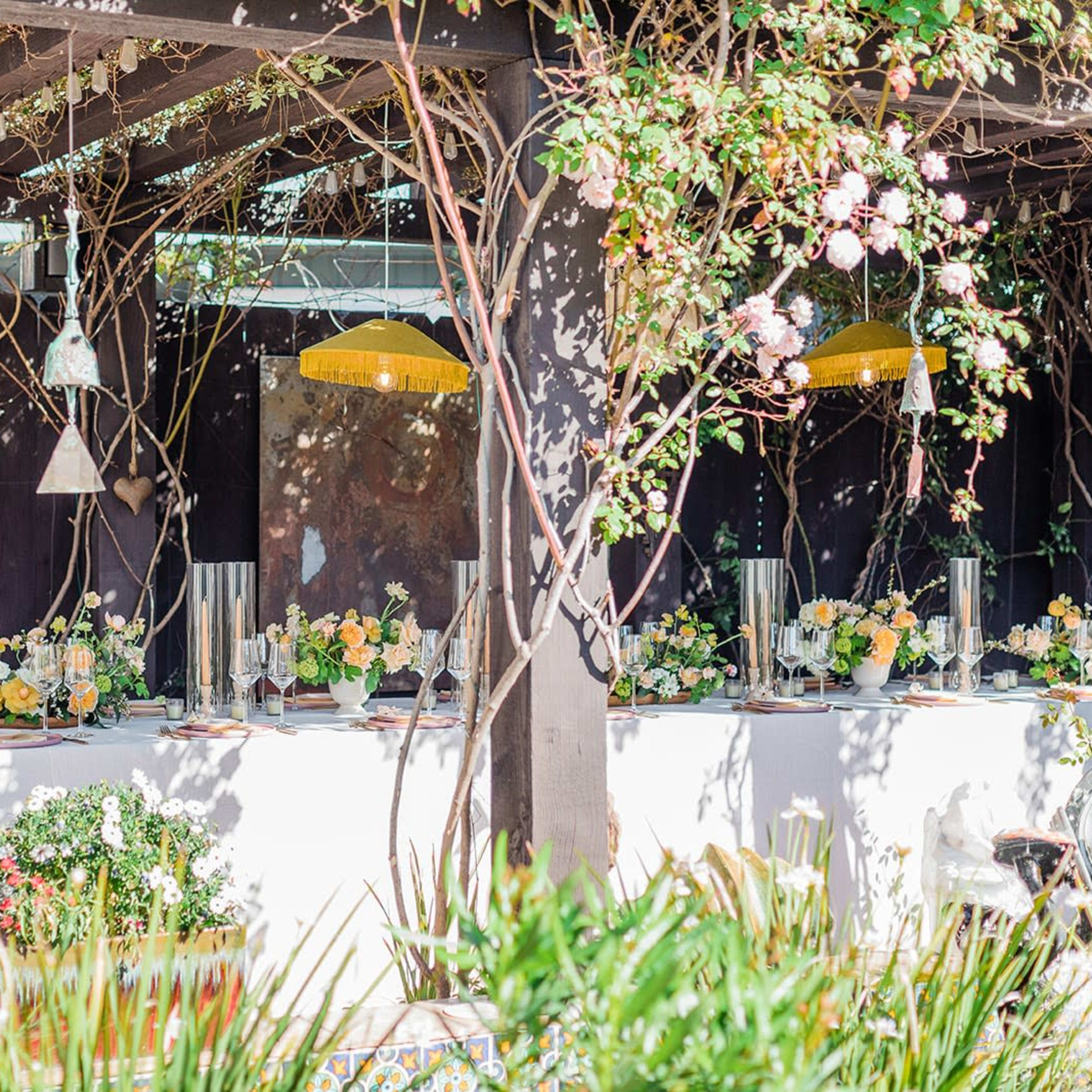 A decorated outdoor dining area features a long table set with floral arrangements and glassware under a pergola adorned with vines and hanging fixtures.