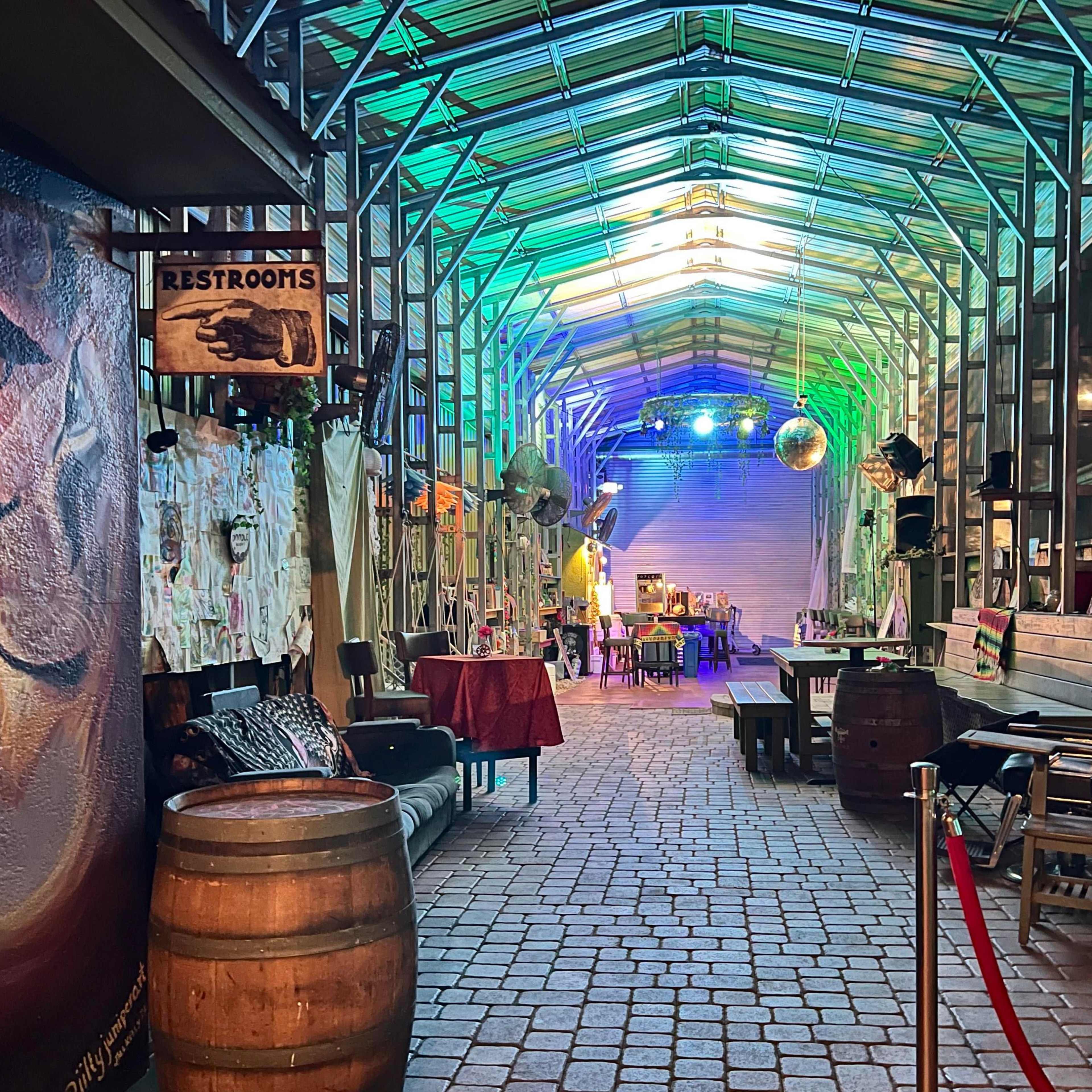 A brightly lit corridor lined with benches and decorative elements, featuring a barrell at one end and a sign for restrooms.