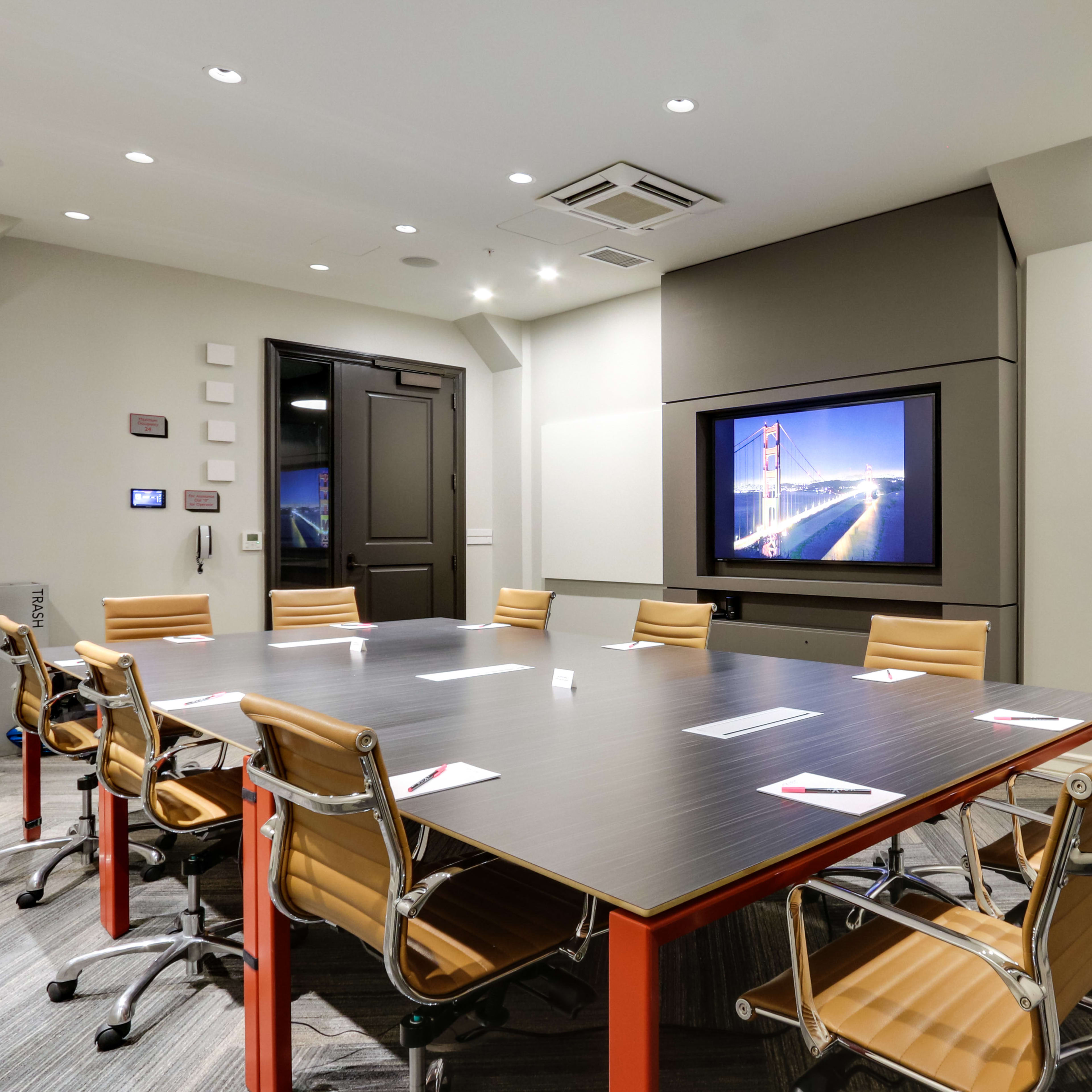 A modern conference room features a large rectangular table surrounded by ergonomic chairs, with a wall-mounted screen displaying an image.