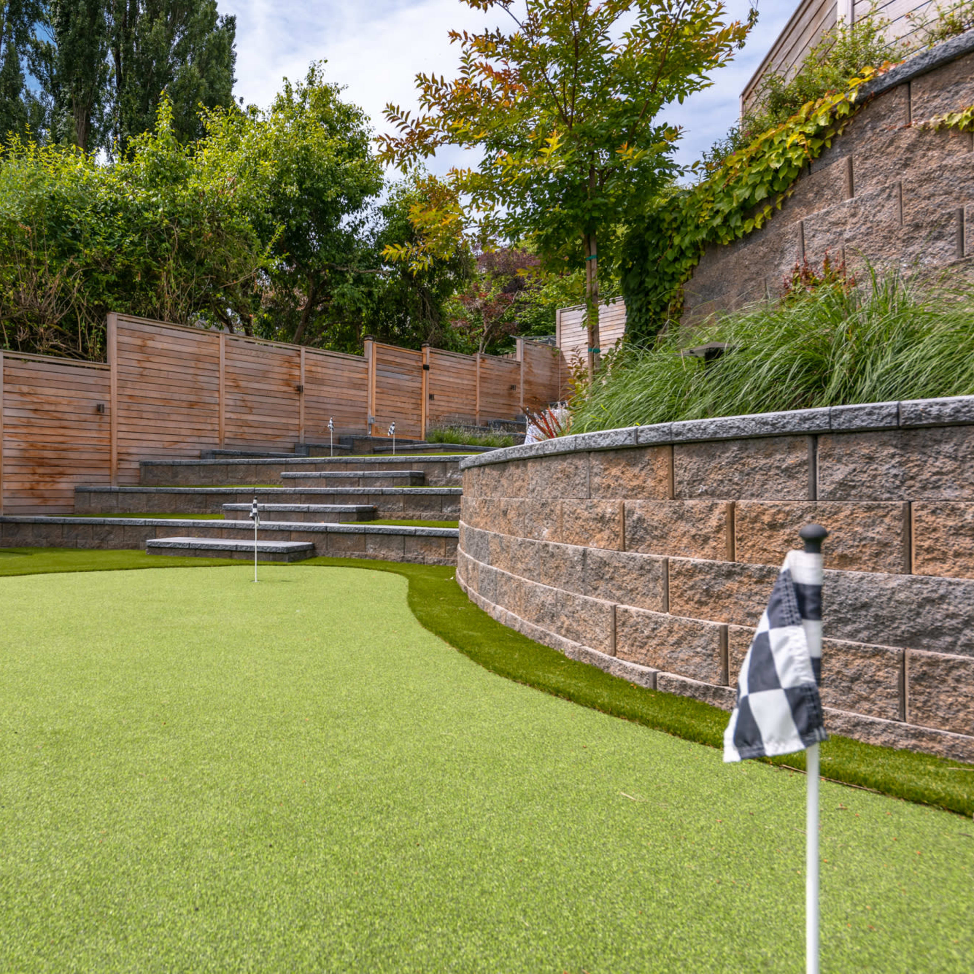 A landscaped putting green with a series of steps and wooden fencing, surrounded by greenery.