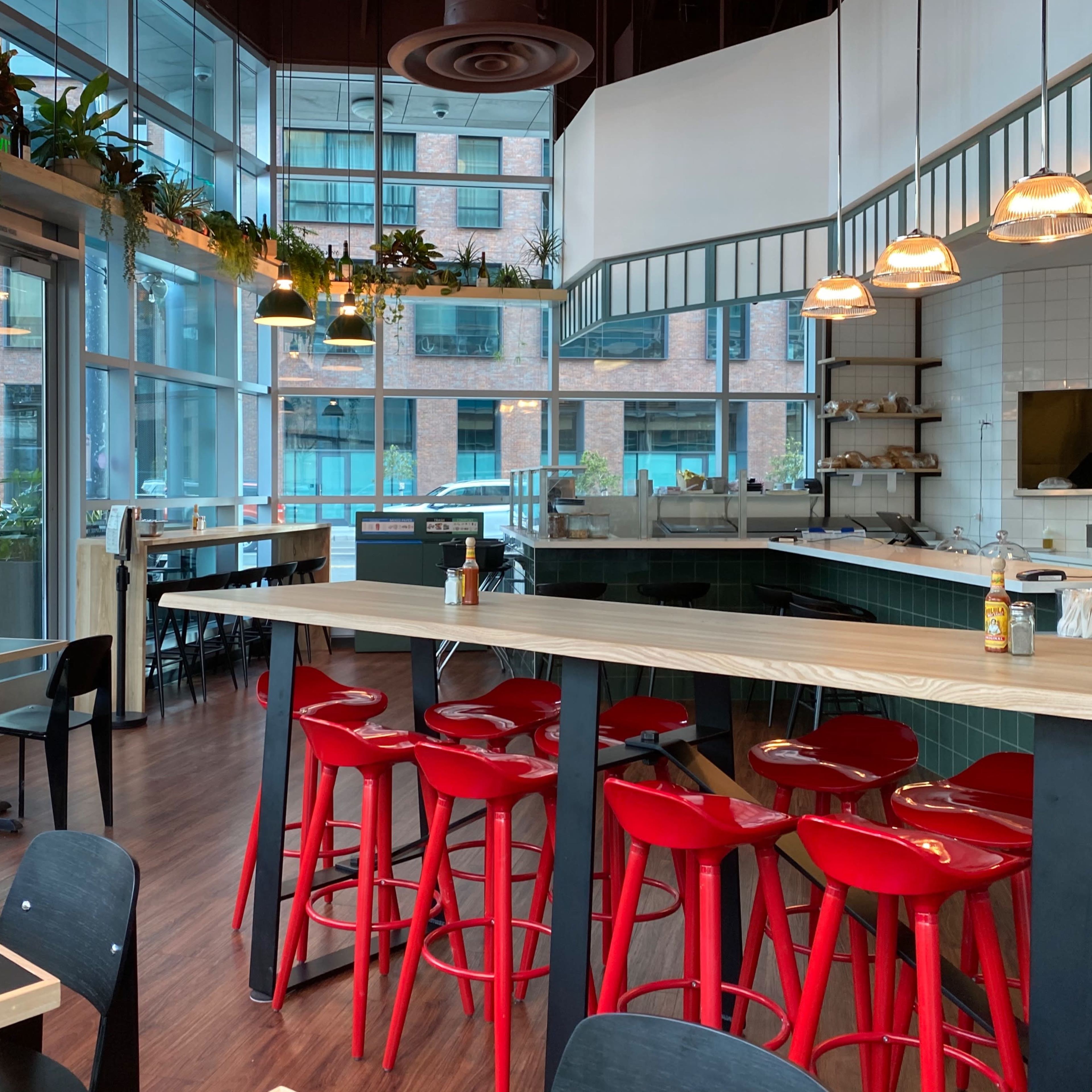 A modern café interior with large windows, wooden tables, red bar stools, and greenery along the walls.
