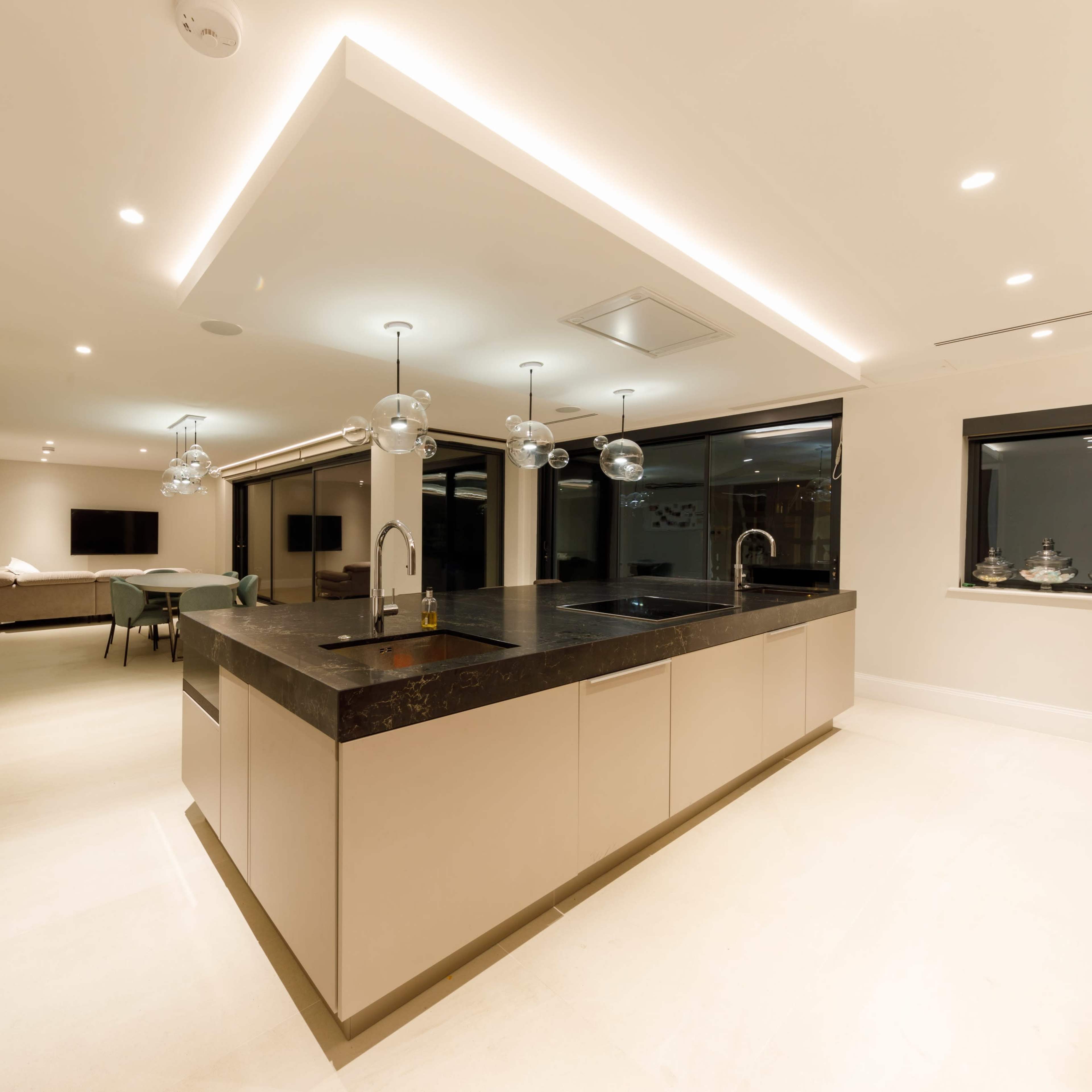 A modern kitchen features a large central island with a dark countertop, sleek cabinetry, and pendant lighting, leading into an open living area with large windows.