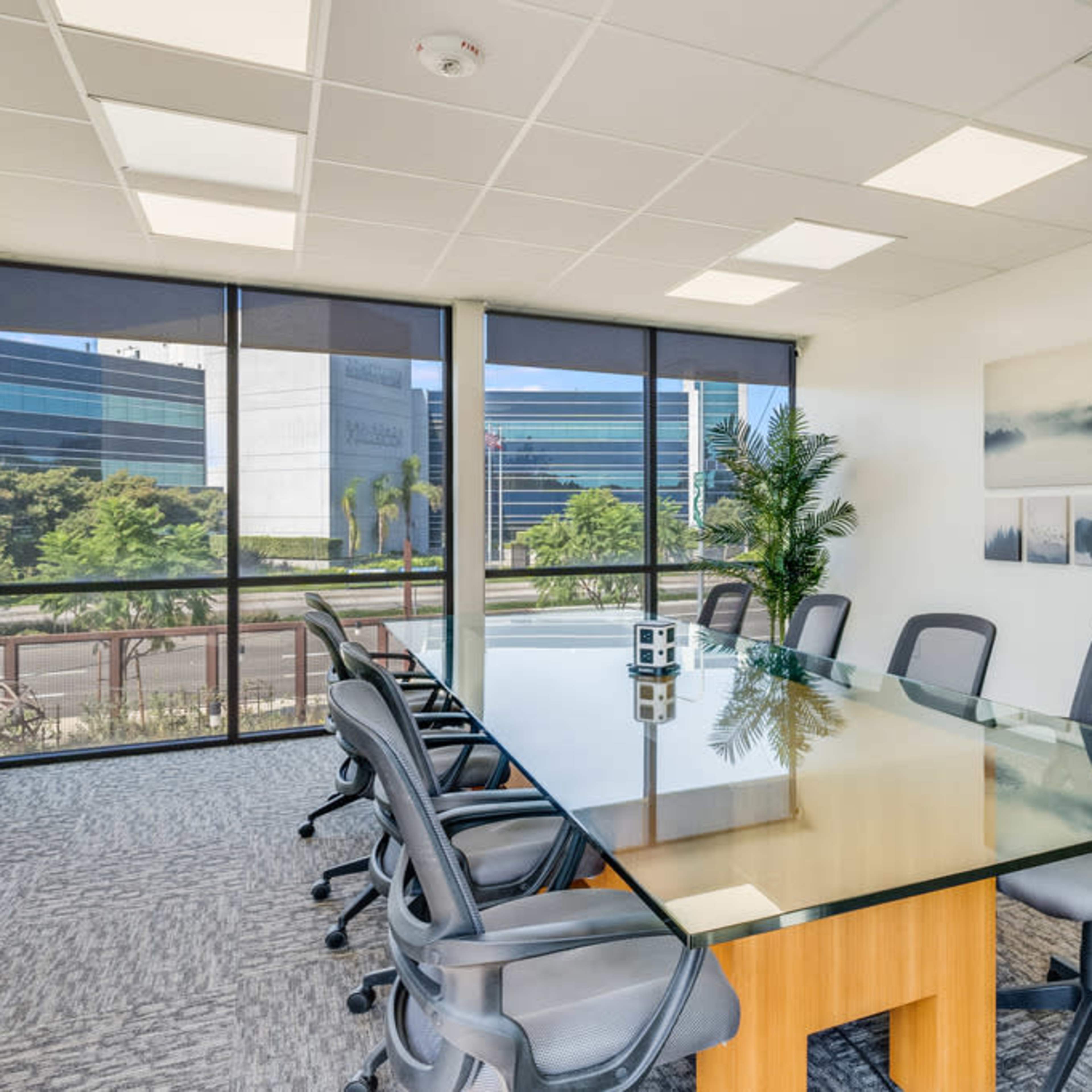 A modern conference room with a large glass table surrounded by several office chairs, and large windows with a view of a landscaped area outside.