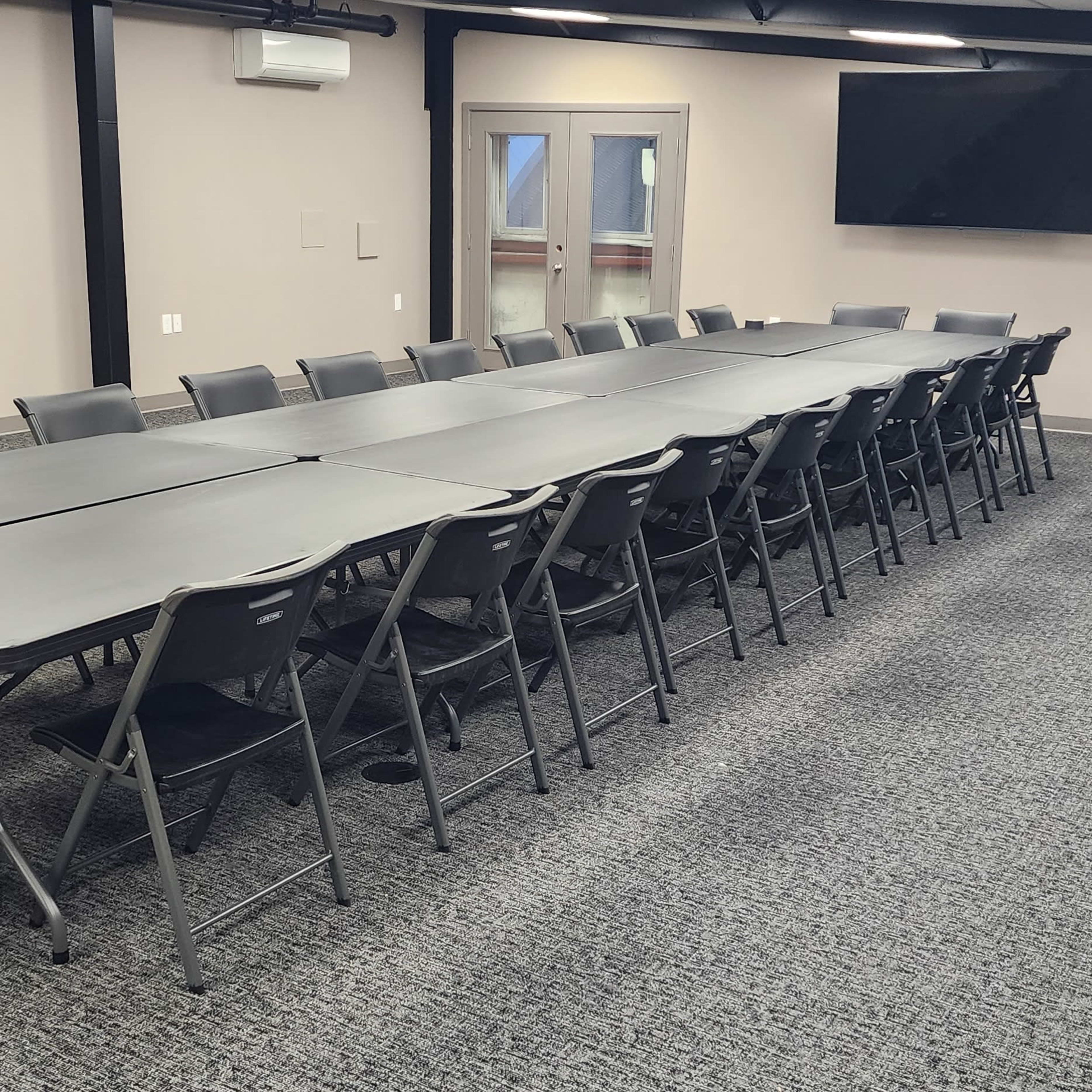 A long arrangement of folding tables and chairs fills a large, empty meeting room with a television and windows along one wall.