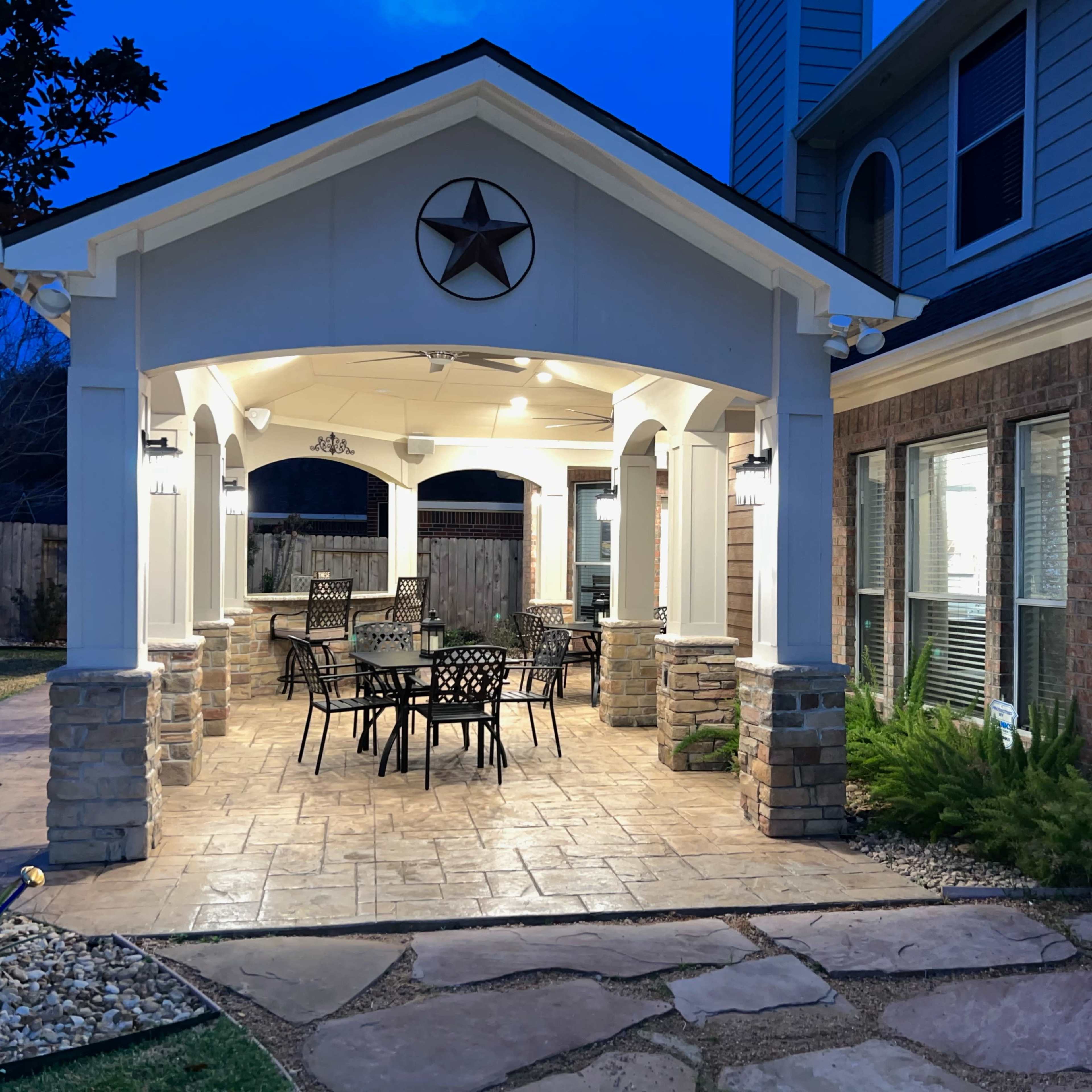 A covered outdoor patio with a dining table and chairs, surrounded by stone flooring and landscaping.