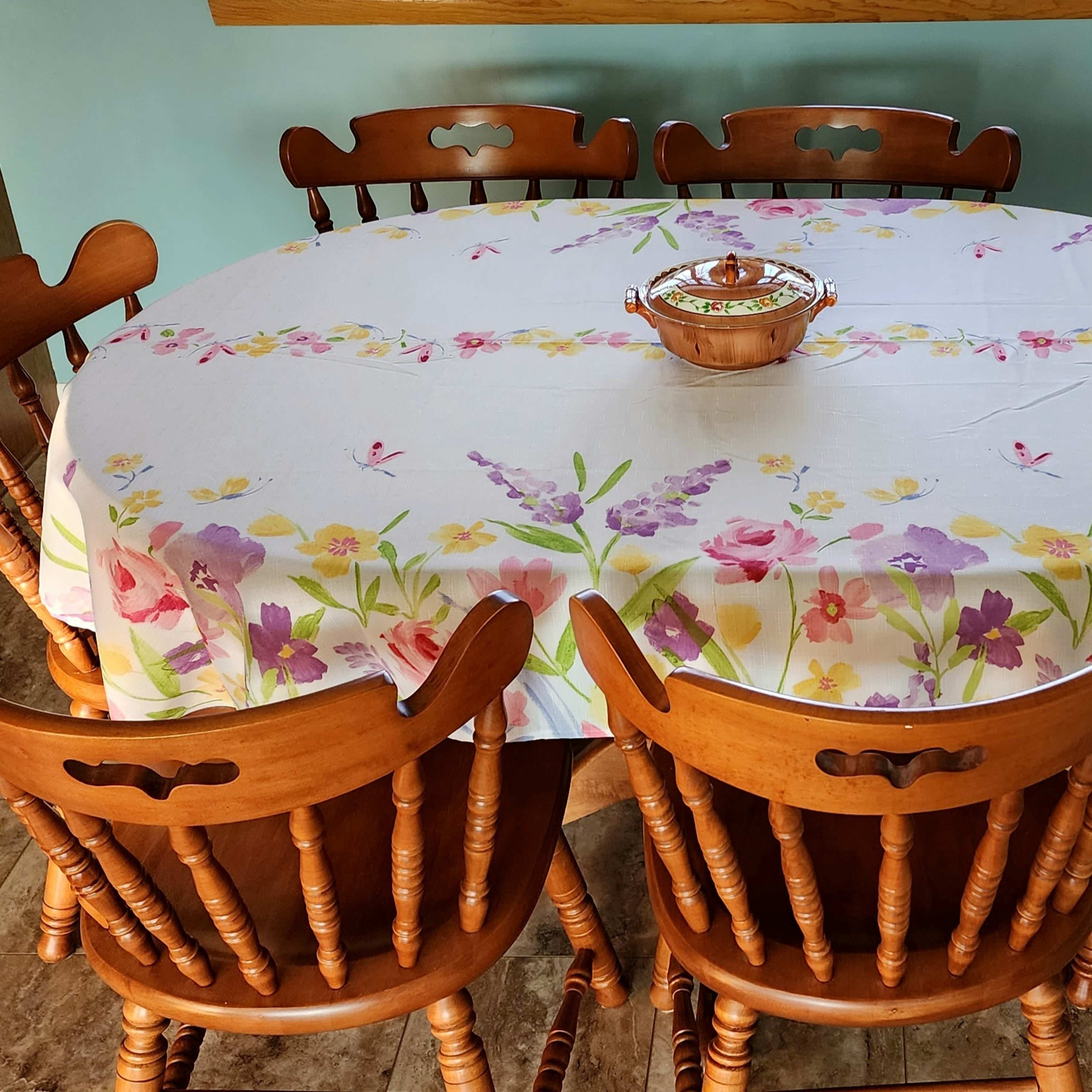 A round wooden dining table with a floral tablecloth and a bowl in the center is surrounded by six wooden chairs.