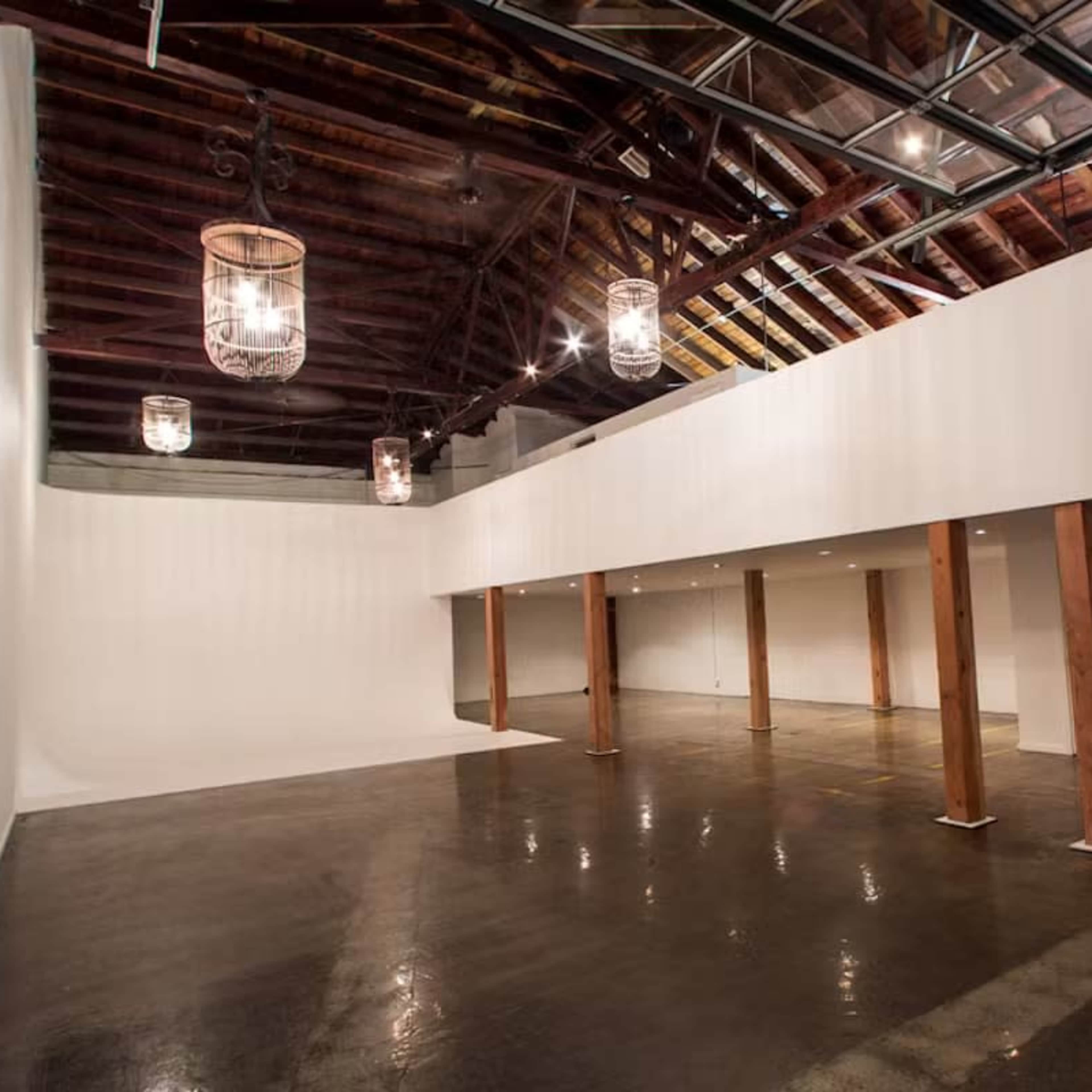 A spacious indoor area with high ceilings, wooden beams, and polished concrete floors, featuring several hanging light fixtures and a large wall for displays or presentations.
