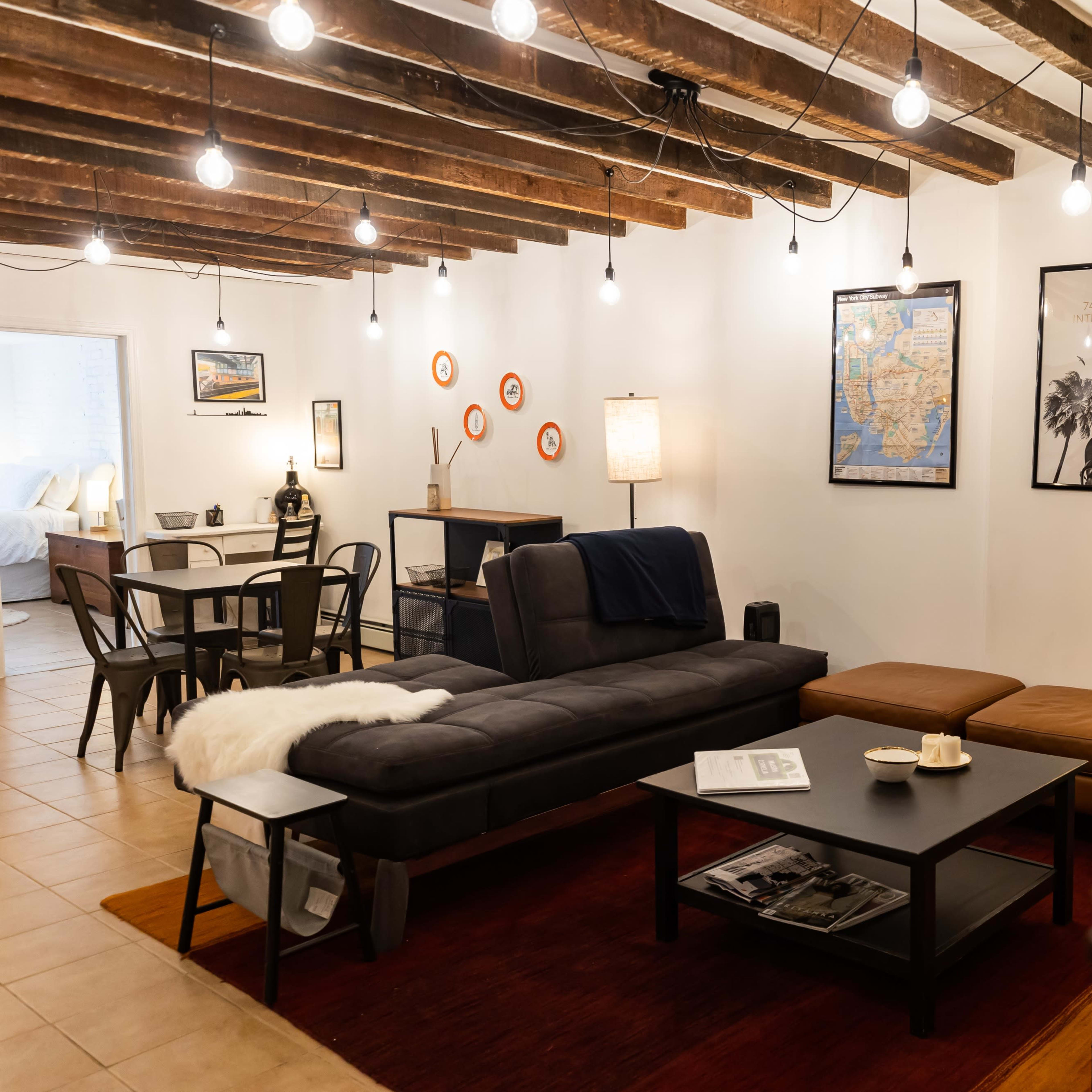 A cozy living room with exposed wooden beams, a dark sofa, an ottoman, a coffee table, and wall decorations including maps and framed pictures.