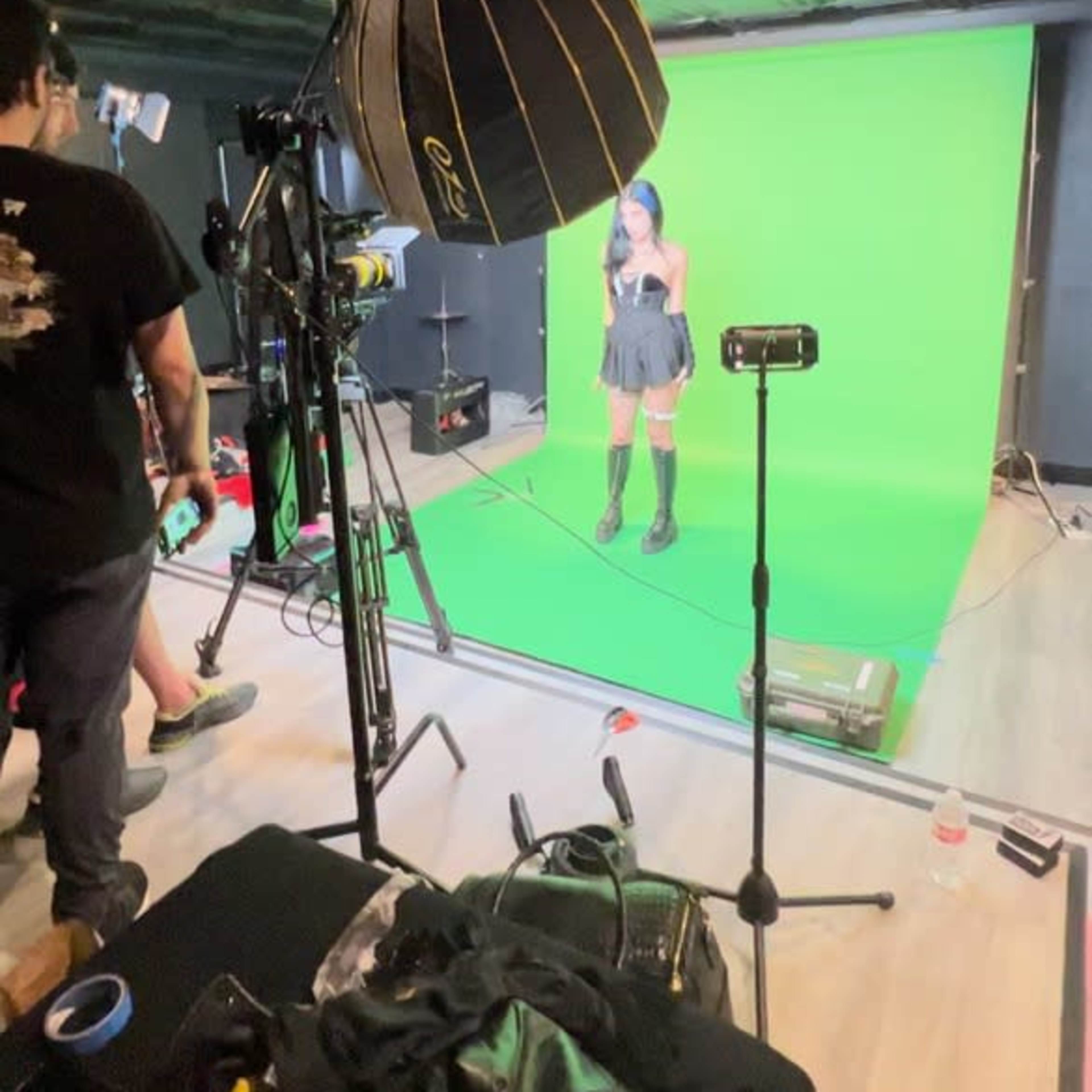 A woman stands in front of a green screen inside a studio, surrounded by cameras and lighting equipment.