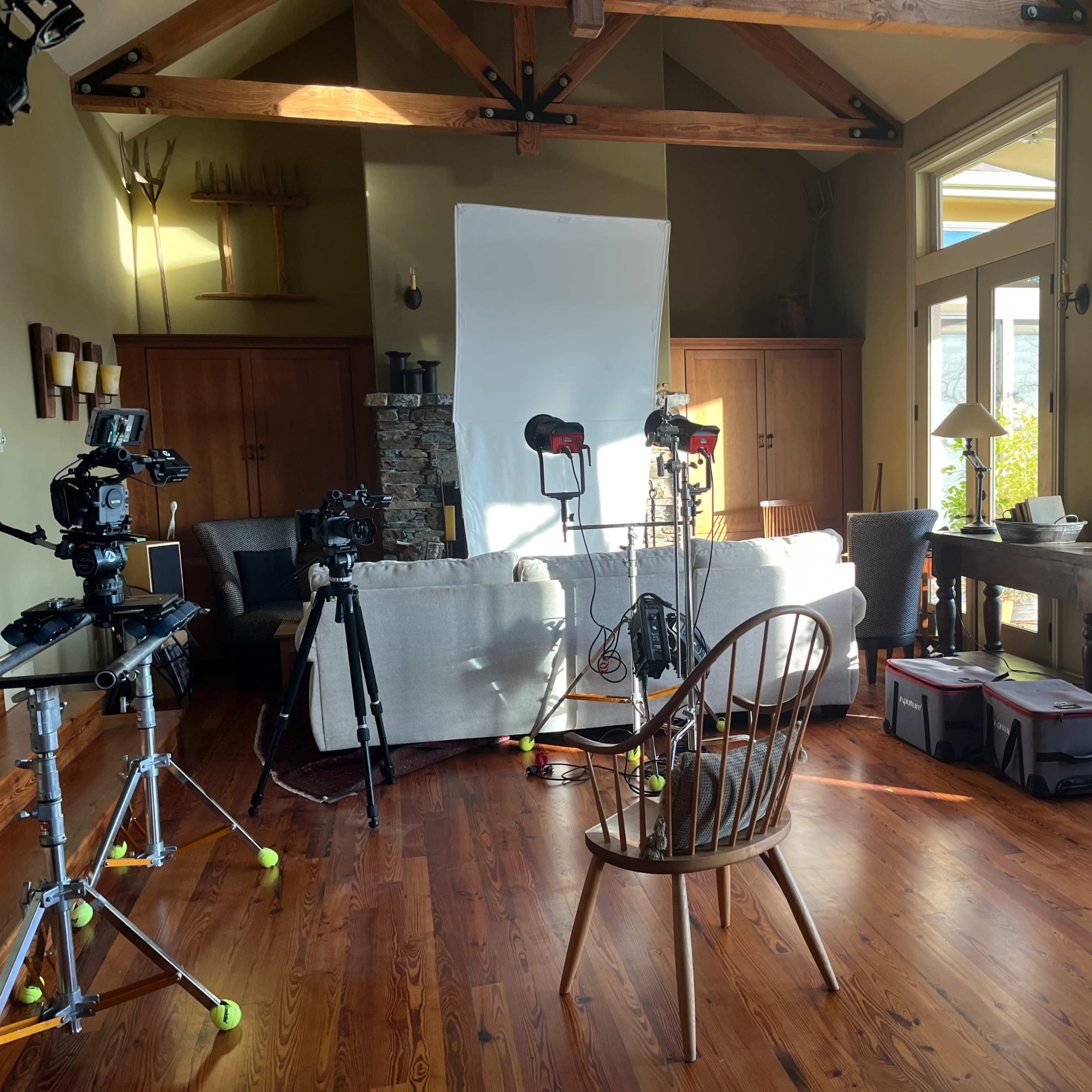 A living room setup for a video shoot, featuring cameras on tripods, a large white reflector, and a wooden chair positioned in the center.