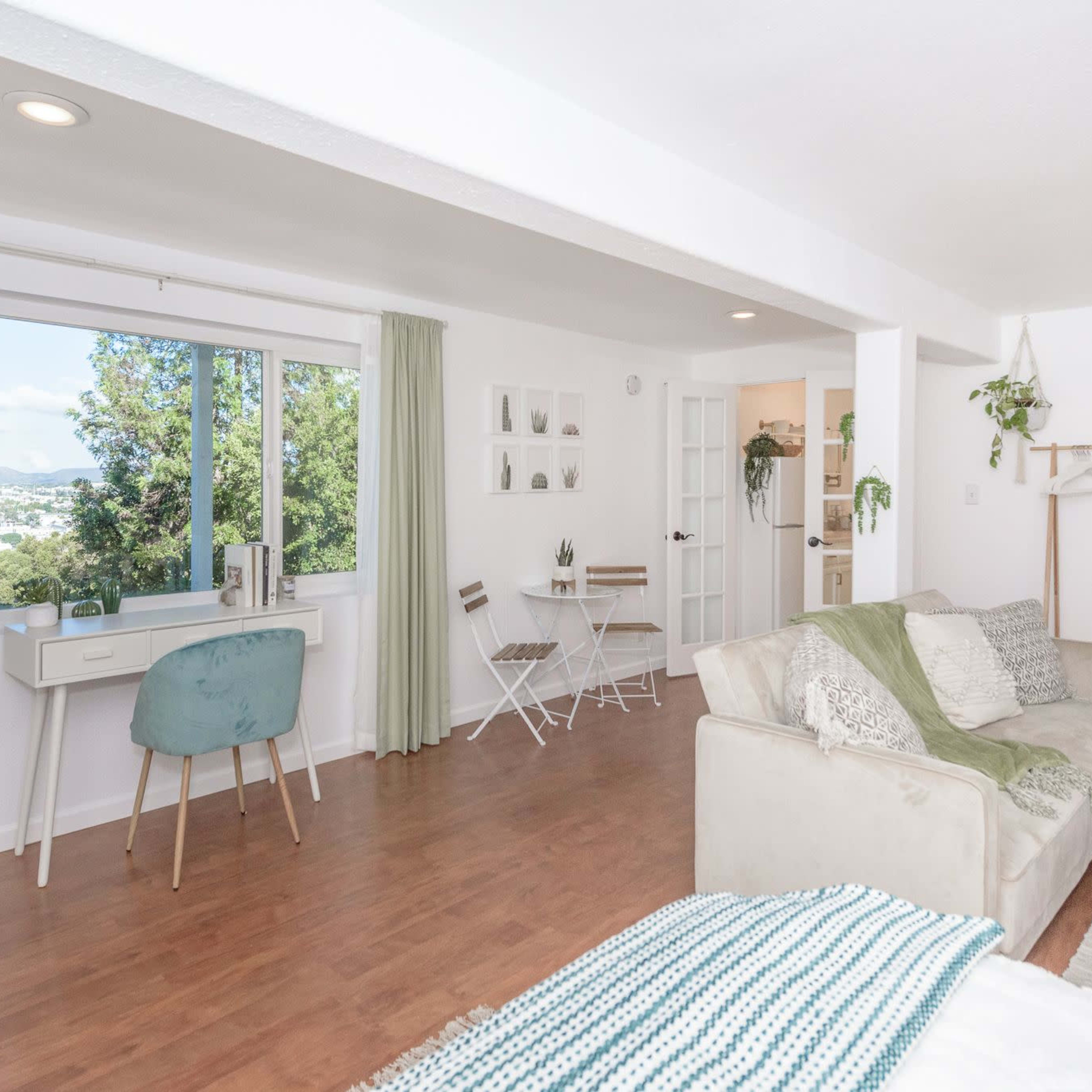 A bright, airy living space features a sofa, a desk by a window, a small dining area with two chairs, and indoor plants.