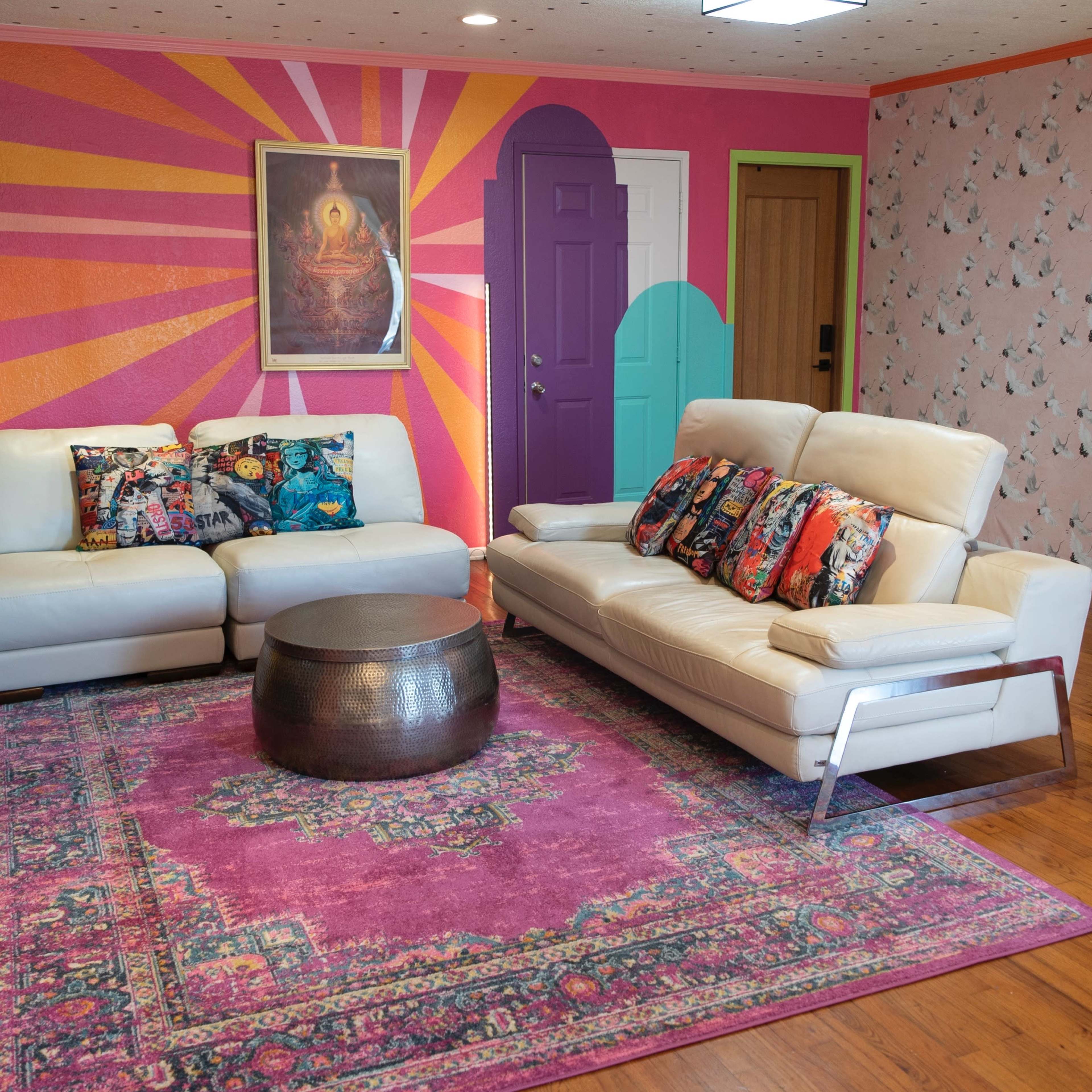 The living room features two white sofas adorned with colorful throw pillows, a wooden table in the center, and vibrant wall decor, including a large mural and patterned wallpaper.