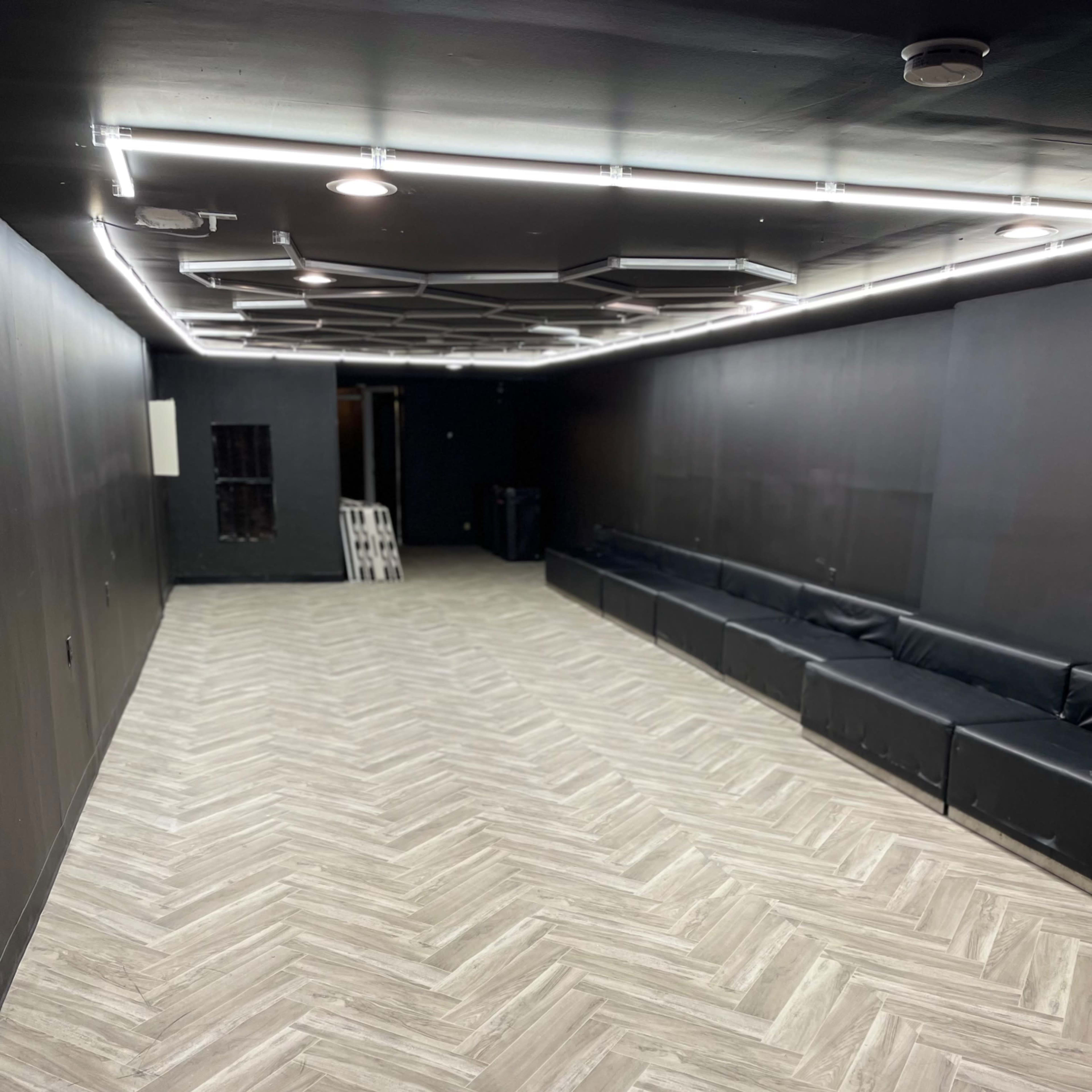 A dimly lit room with herringbone-patterned flooring, black walls, and a long black couch along one side.