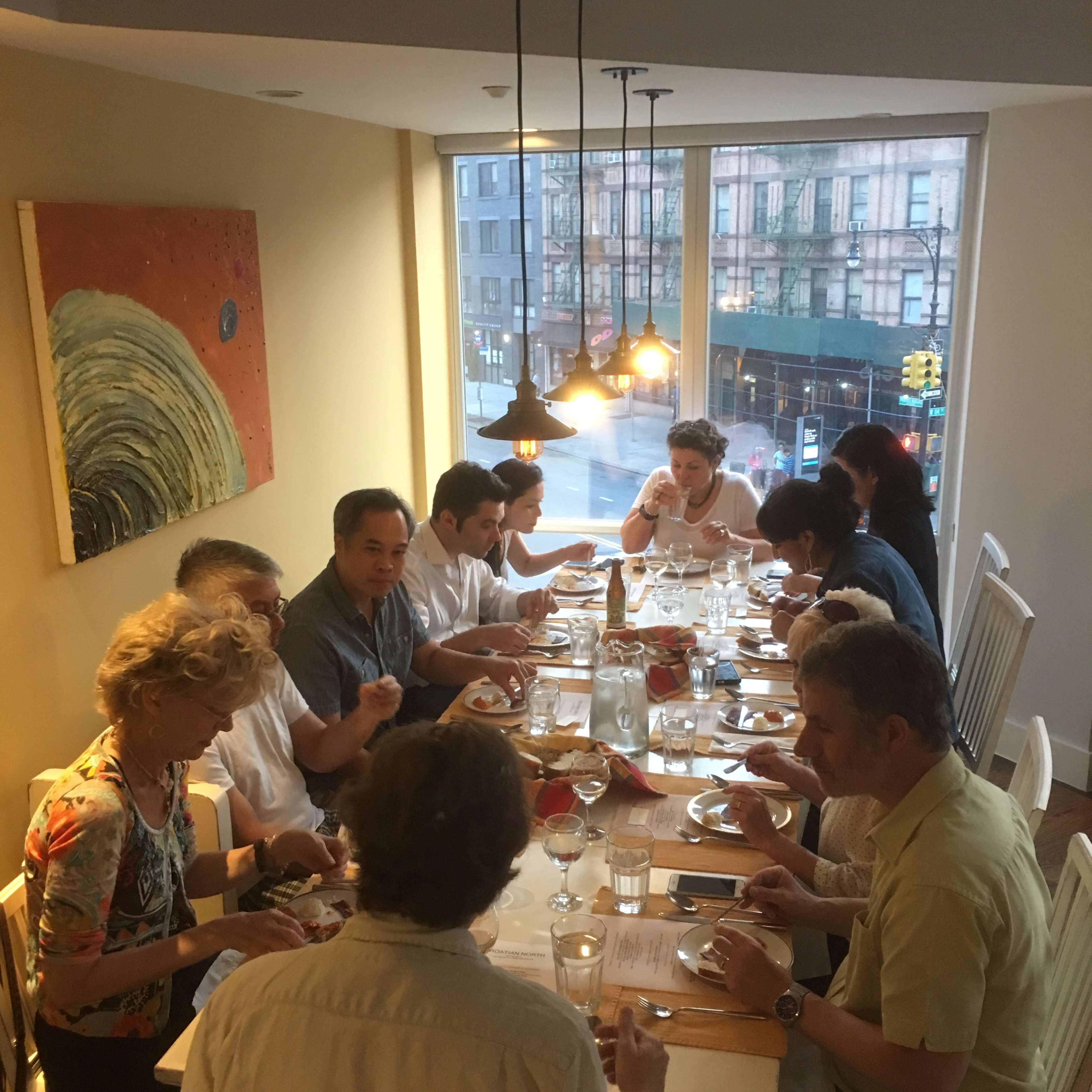 A group of twelve people sits around a long table in a well-lit dining room, enjoying a meal together.