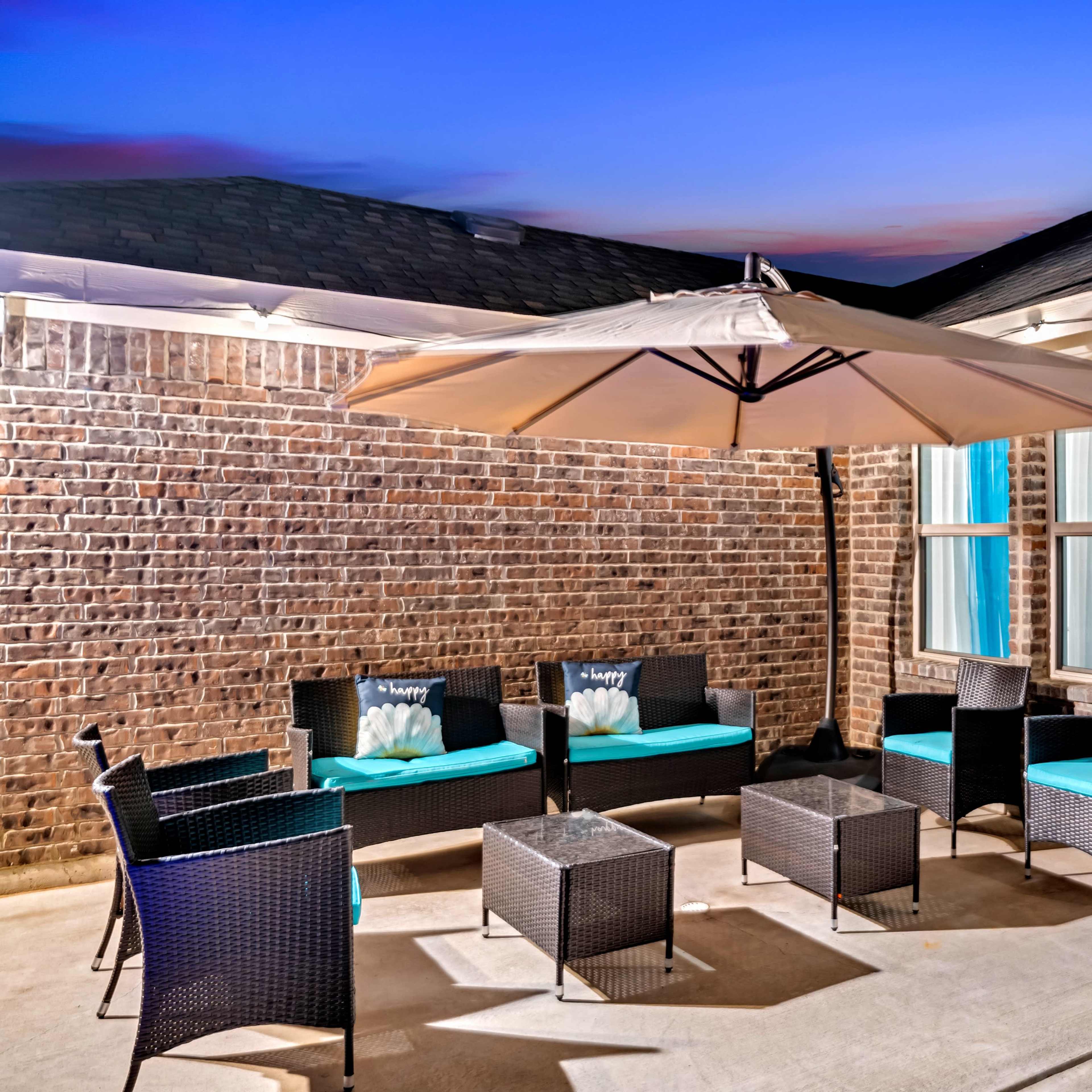A patio area features a large umbrella over a seating arrangement with several chairs and a table against a brick wall.