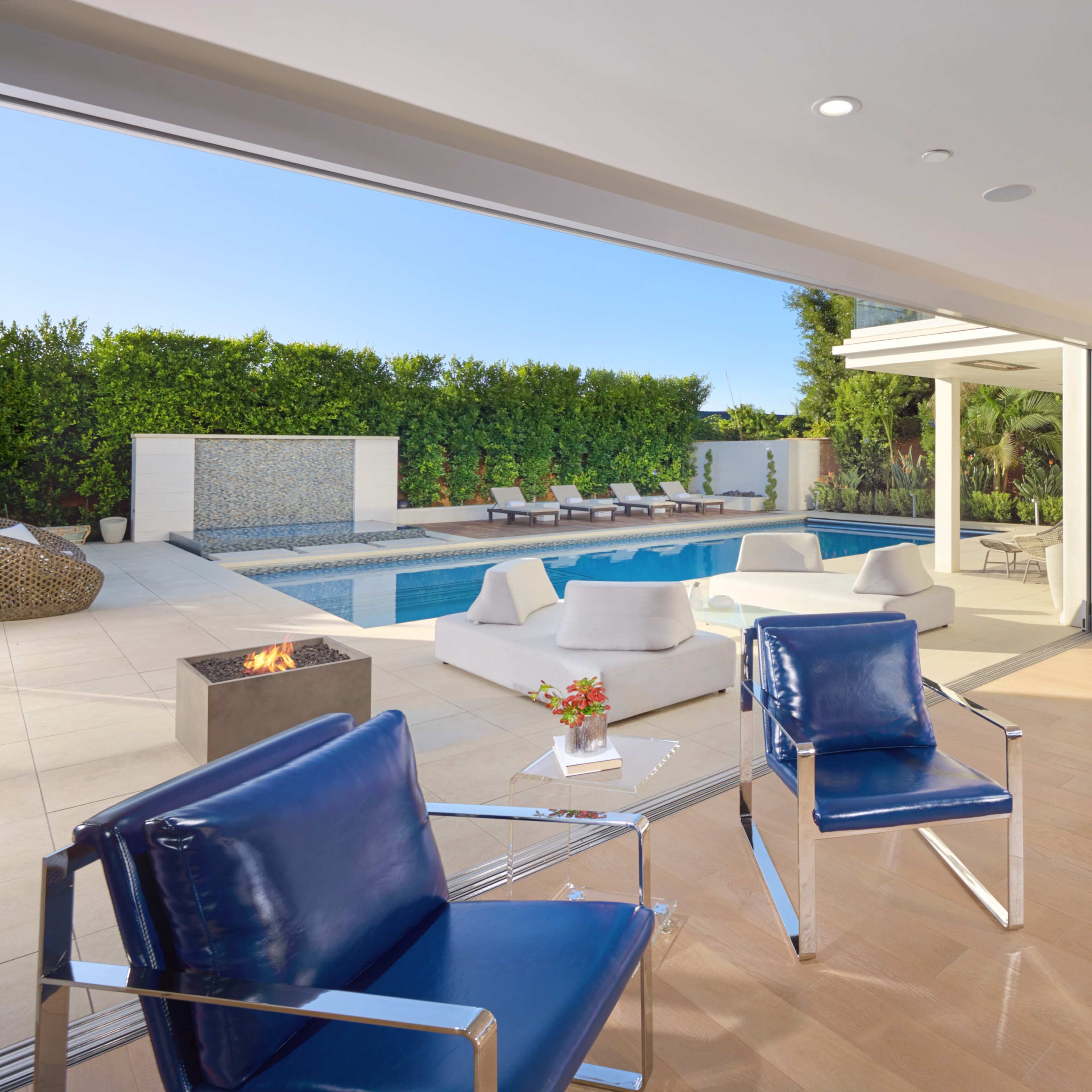 A modern indoor space with blue chairs, opening to a patio with a swimming pool and sun loungers surrounded by greenery.