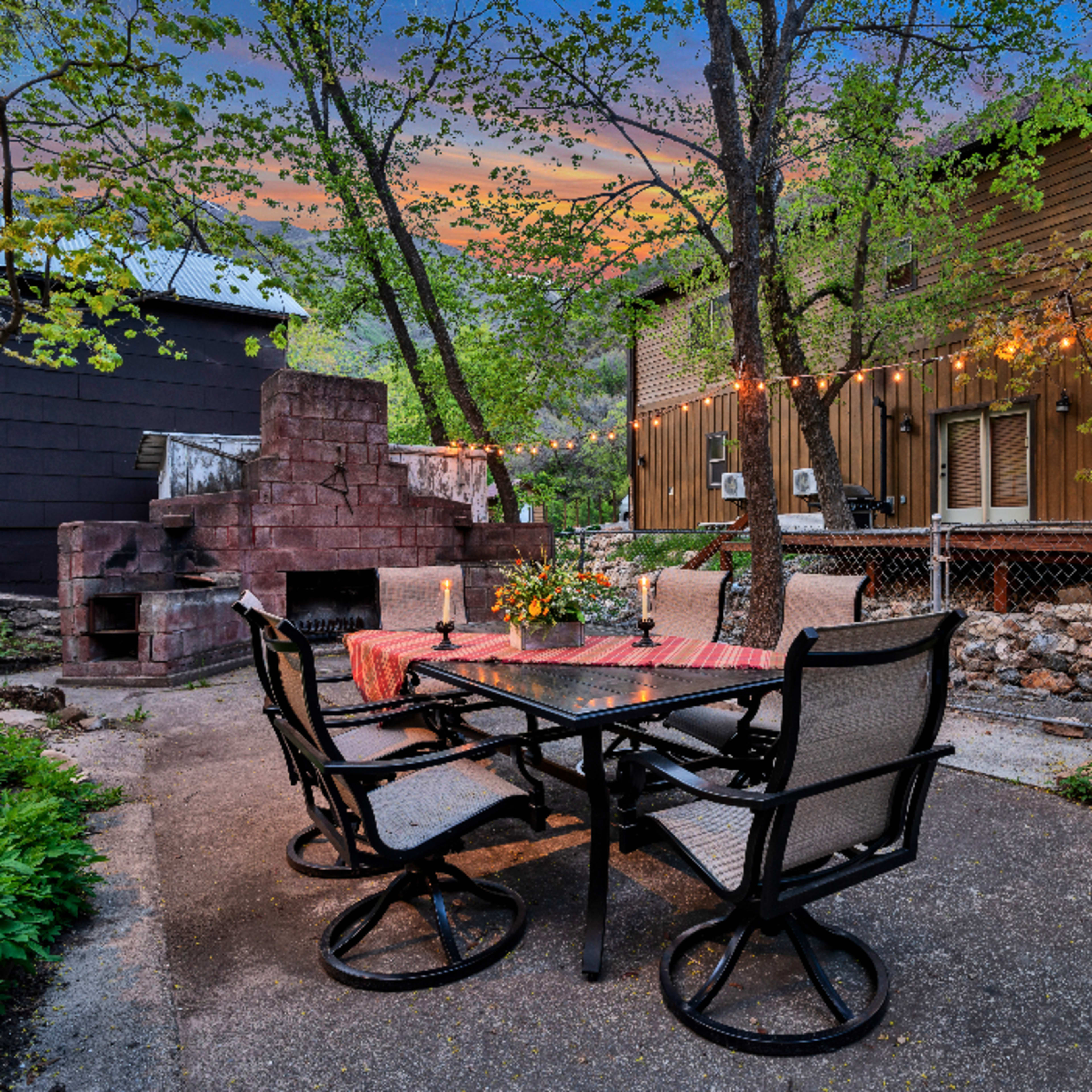 A patio area featuring a dining table with chairs, an outdoor fireplace, and decorative string lights, surrounded by trees and buildings.