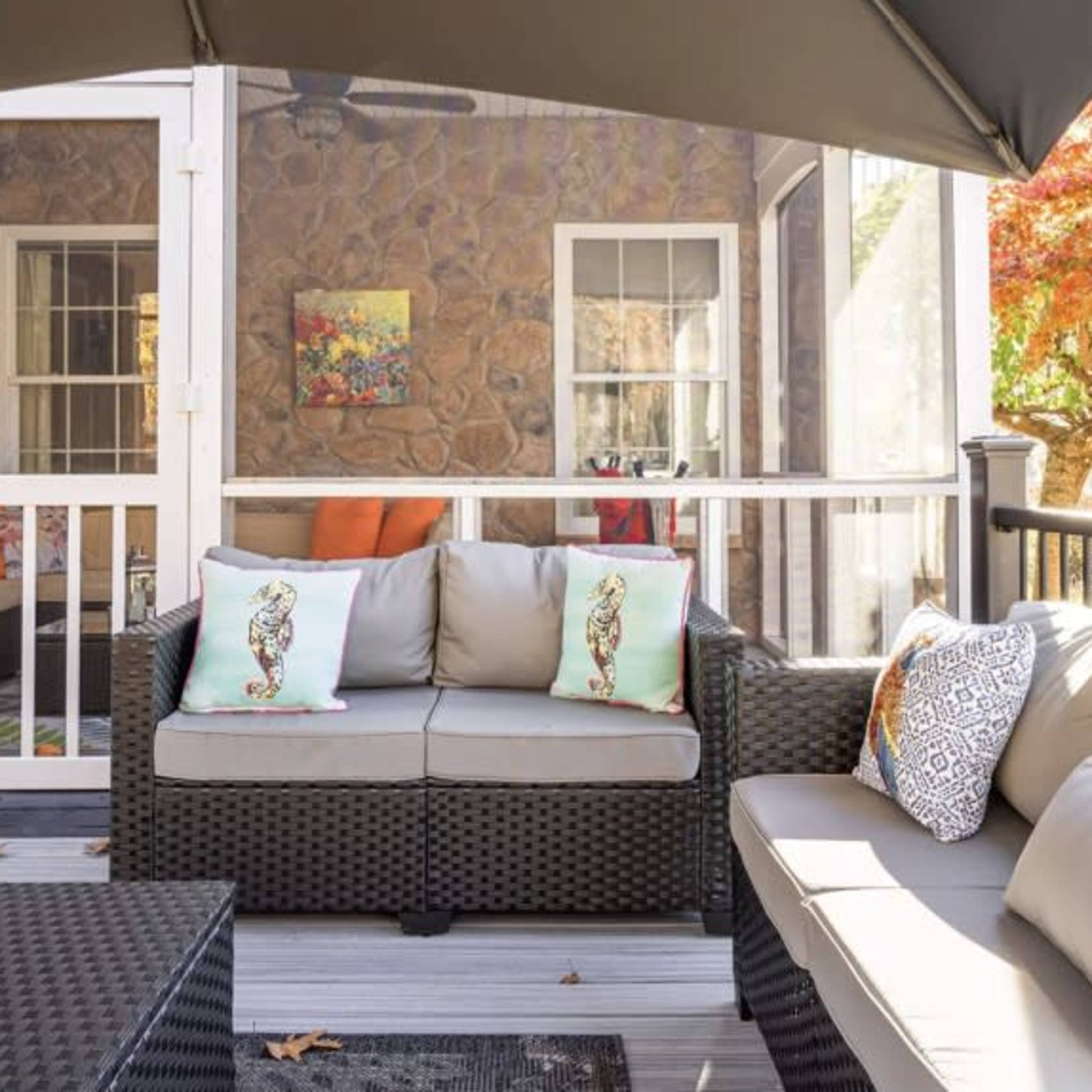 A screened porch features a seating area with gray sofas adorned with decorative pillows, overlooking a stone wall and an autumn tree outside.
