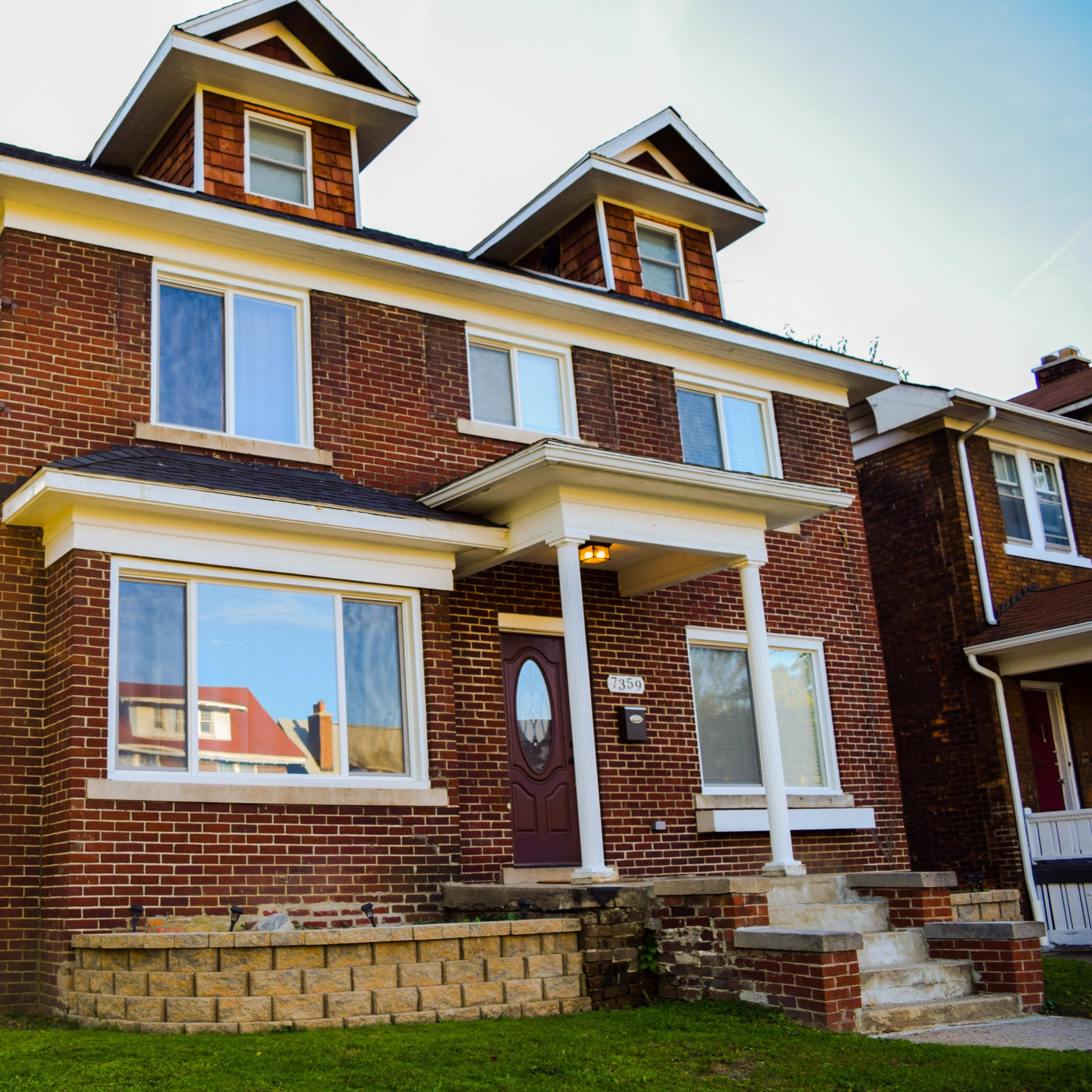 A red brick house with two dormer windows and a large front porch is situated on a grassy lawn.