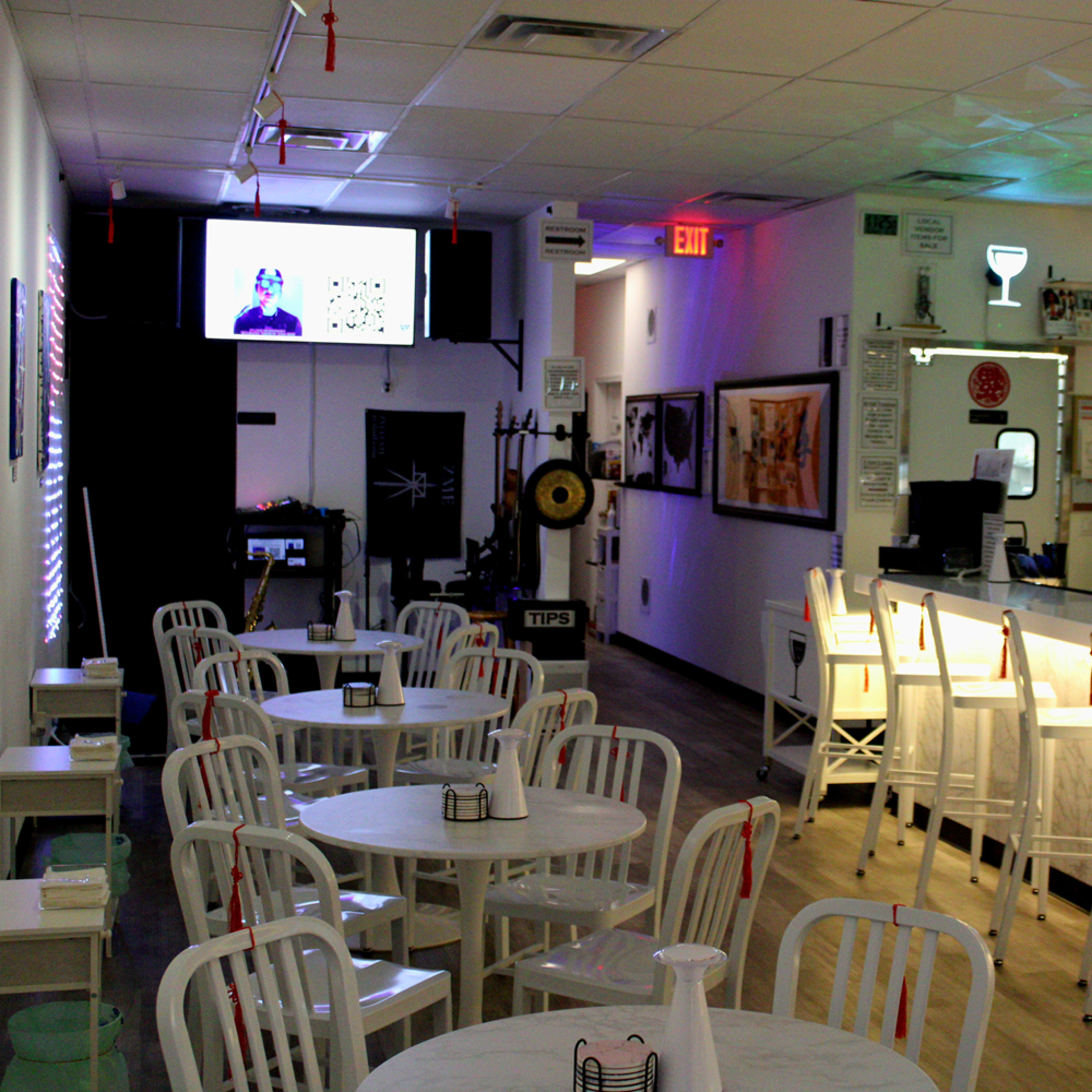 A modern restaurant with white tables and chairs, a bar area with illuminated shelves, and a large screen displaying video content.